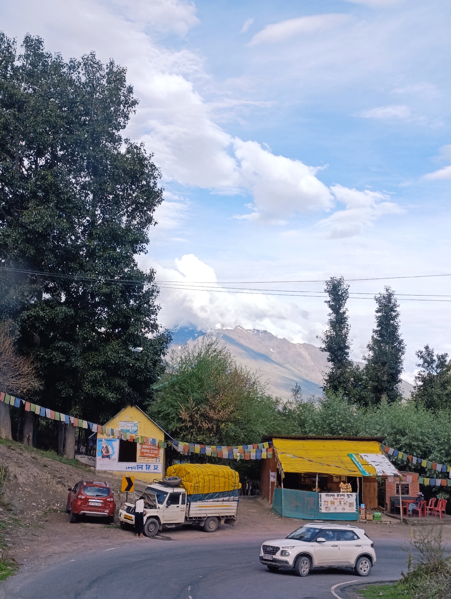 In Gondhla, India, this outdoor scene depicts a roadside with several elements. A large, dark green tree with dense foliage dominates the left side of the frame, partially obscuring a cloudy sky with blue patches. In front of the tree, a small yellow building with a blue-green awning stands, adorned with colorful prayer flags. To its left, a red SUV is parked. Next to the yellow building, a white pickup truck with a yellow-covered cargo bed is parked. A person, possibly male, is standing near the open hood of the truck. Further to the right, another small building with a yellow roof and orange walls serves as a roadside stall. Red chairs are set up outside this stall. A white SUV is driving on the paved road in the foreground. In the background, a majestic mountain range with snow-capped peaks is visible, partially shrouded by clouds and tall trees. The overall impression is a rural, mountainous landscape with signs of local commerce and transportation.