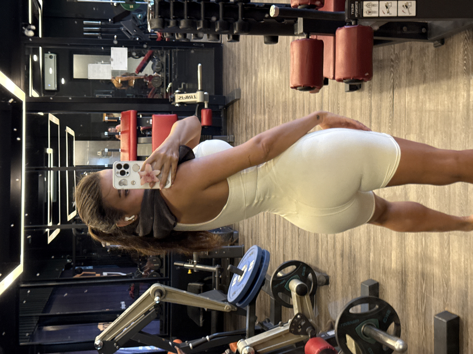 A woman wearing a white athletic outfit is taking a selfie in a gym. She holds a smartphone with a white case adorned with a floral design. She is wearing a grey towel and white wireless earbuds. The gym contains weightlifting machines, barbells, and a leg press machine. The background shows mirrors reflecting other gym equipment. The photo appears to be taken in North Kuta, Indonesia, based on information provided.