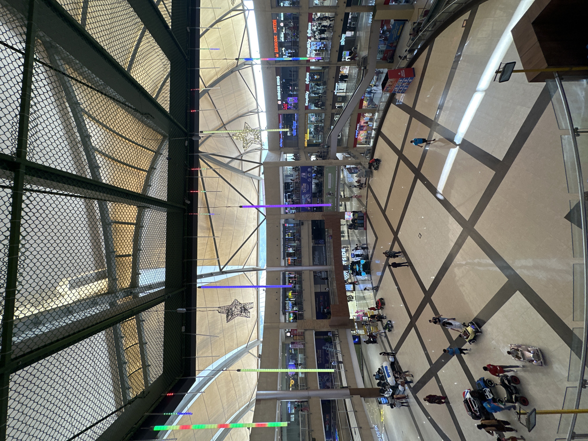 Here's a detailed description of the image:
The image shows the interior of a multi-level shopping mall in Kalyan-Dombivli, India, viewed from an elevated position looking down into the central atrium. The atrium floor is tiled with a patterned design. Several people can be seen walking around and operating ride-on toy cars.
The surrounding levels of the mall are lined with shops and restaurants. Signage for various businesses such as "Burger King," "Easy Buy," and "Barbeque Nation" are visible. There are lights suspended from the ceiling, some arranged in vertical lines, and a decorative star-shaped light fixture.
A section of the view is obstructed by a wire mesh, suggesting that the photograph was taken from behind a protective barrier, possibly a balcony or a high walkway.