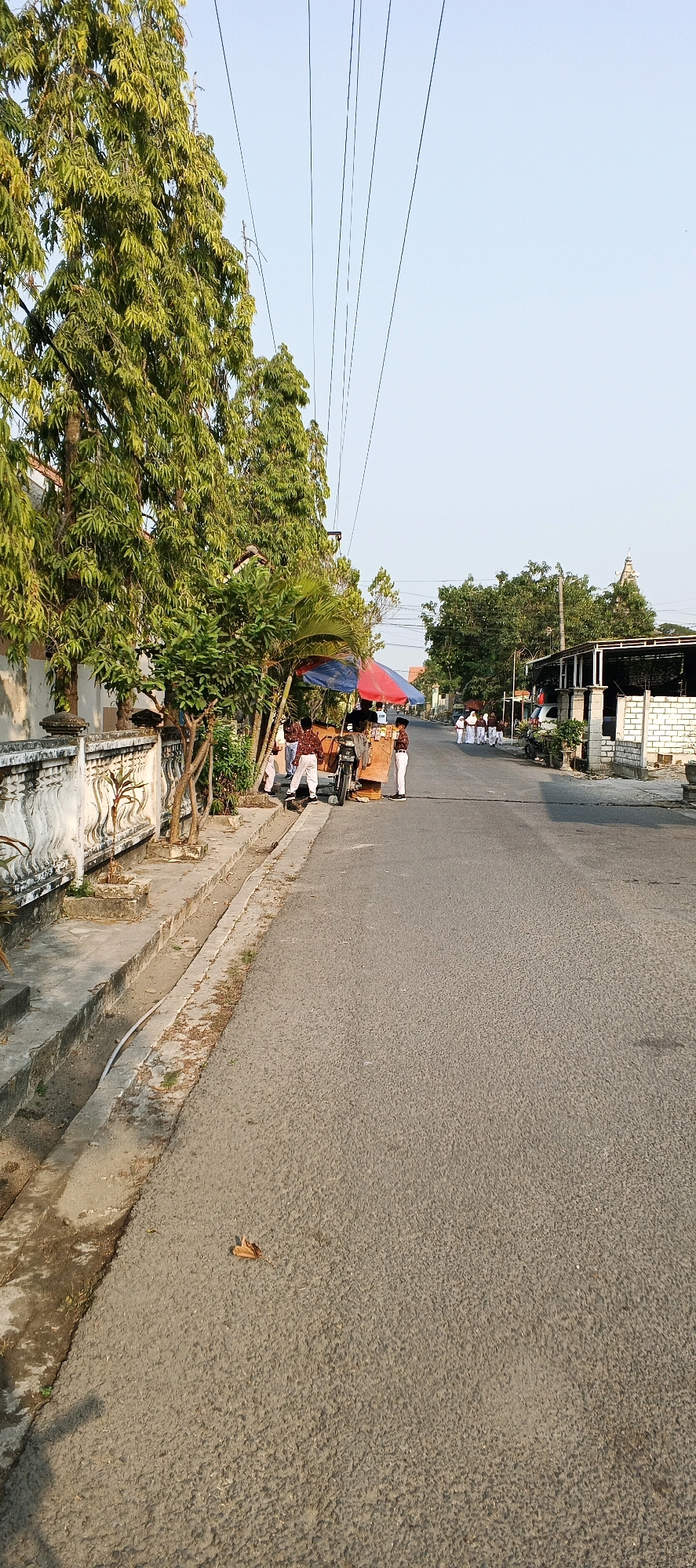 The image depicts a street scene in Latukan, Indonesia, with a group of people, possibly students in uniform, gathered near a food cart under a colorful umbrella. Tall green trees line the left side of the street, which appears to be paved. A white decorative wall runs along the side of the road. In the distance, more buildings and trees are visible, and power lines stretch across the sky. The lighting suggests it is daytime, with clear sky.