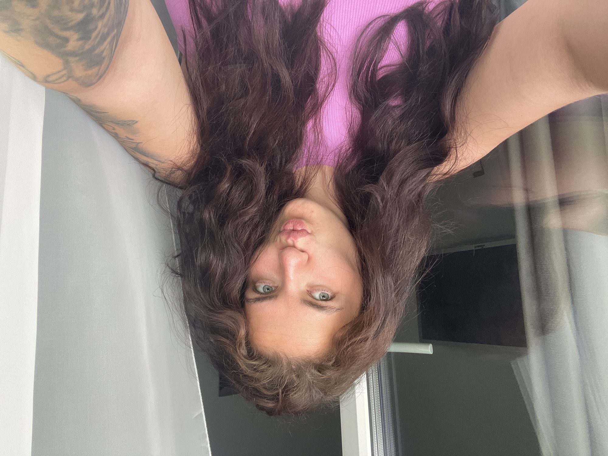 A woman with long, dark, wavy hair is depicted in a selfie taken from an upside-down perspective. She is wearing a pink tank top. Her face is centrally framed, and she has light eyes and pursed lips. A tattoo is visible on her left arm. A white curtain hangs on the left side of the frame, and a window is seen to the right. The location is Мосметаллоконструкция Онт, Russia.