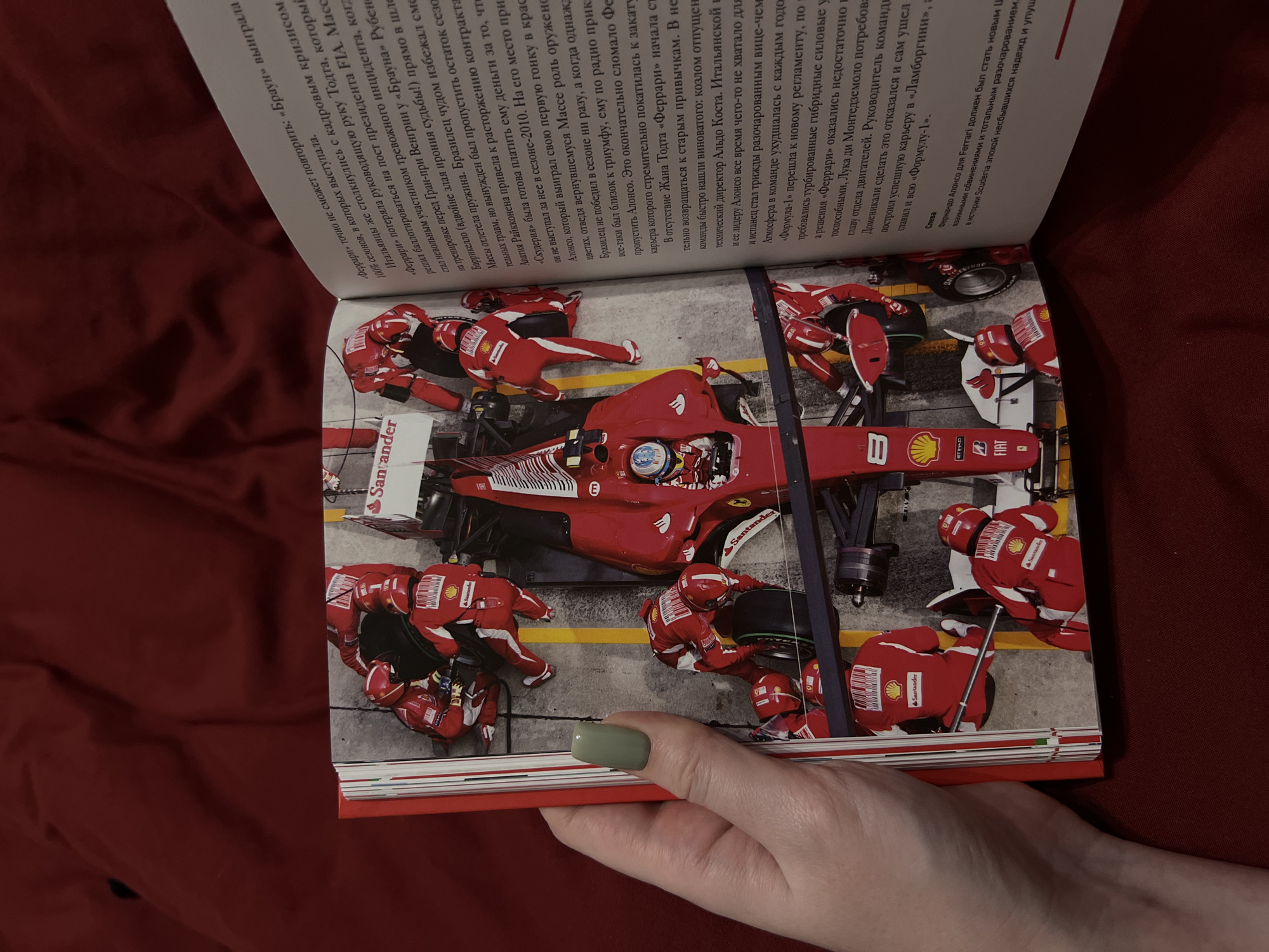 A human hand with a light green fingernail is holding open a book displaying a photograph of a Formula 1 pit stop. The photograph shows a red Ferrari race car surrounded by mechanics in red racing suits. The mechanics are in the process of changing a tire. The car has the "Santander" logo visible on it. The text on the right page discusses Ferrari and its history. The background is dark and indistinct. There is no information about the city of Дачное, Russia in the image or accompanying text.