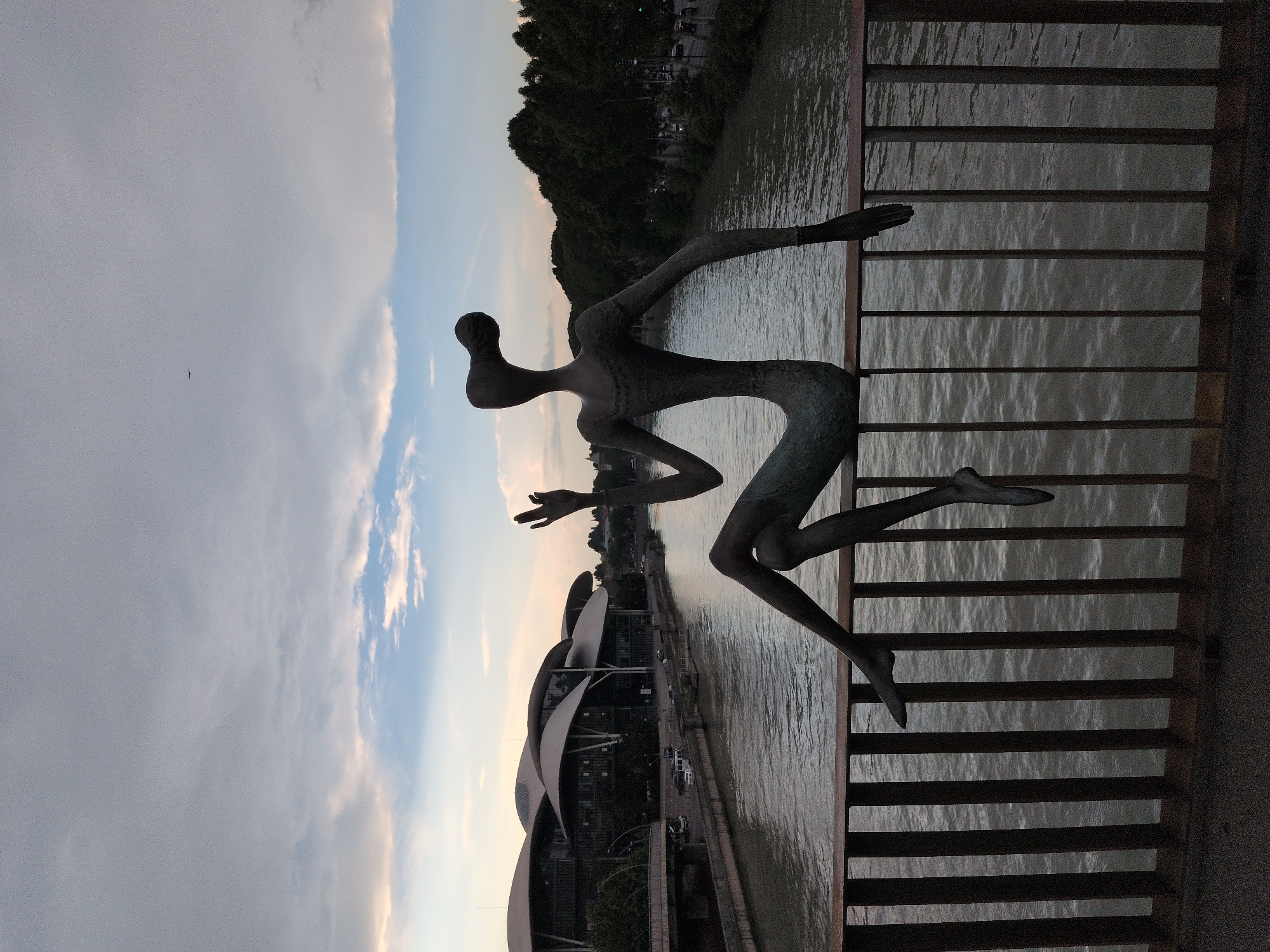 A bronze sculpture of a seated figure with elongated limbs and an abstract head is positioned on top of a dark metal railing, which spans across a body of water. The figure is seen from the back, with its right arm raised in a gesture of salutation or attention and its left arm resting by its side. Its legs are bent, with the left leg dangling over the railing and the right leg tucked underneath. The sculpture is situated on the Peace Bridge, a pedestrian bridge in Tbilisi, Georgia, which crosses the Kura River. In the background, across the river, are modern buildings with distinctive curved roofs, the Parliament of Georgia building, and a tree-lined embankment. The sky above is overcast with some patches of light blue and soft clouds, suggesting either early morning or late evening.