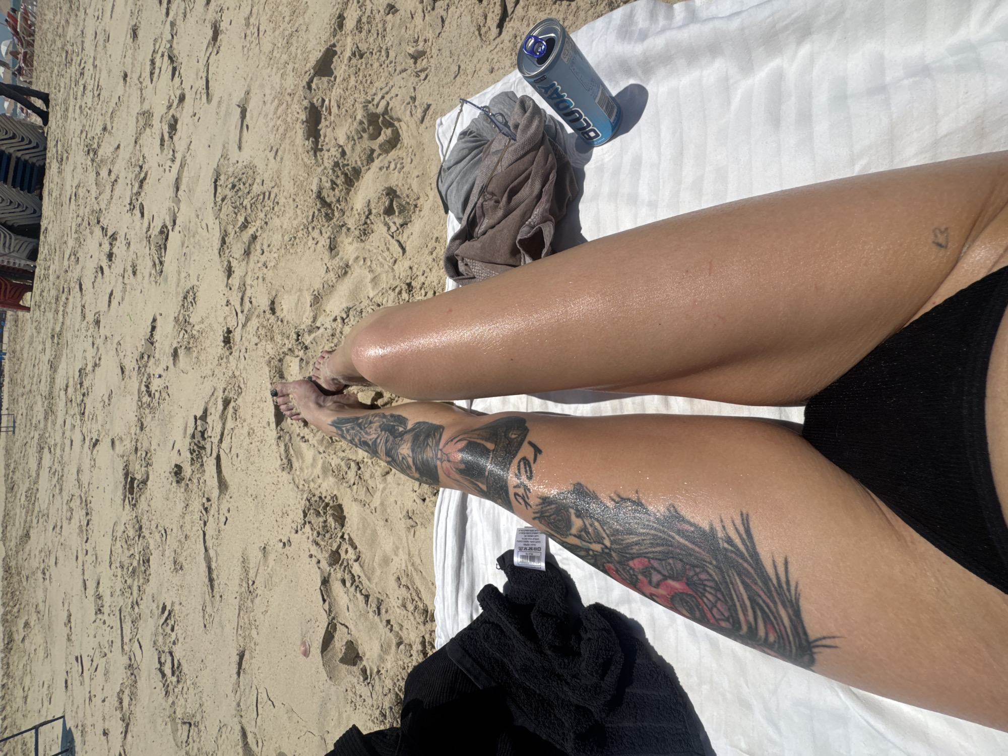 A person with tattooed legs lies on a white towel on a sandy beach. The tattoos are visible on the right leg, one prominently featuring a woman's face and a feather design. A partially consumed can of "BLUODY" sits near a pile of clothing, alongside a pair of sunglasses. The beach is sandy, and beach chairs are visible in the background. The location is suggested as Bat Yam, Israel. The person is wearing black swimwear. The legs are tanned and possibly oiled or wet.