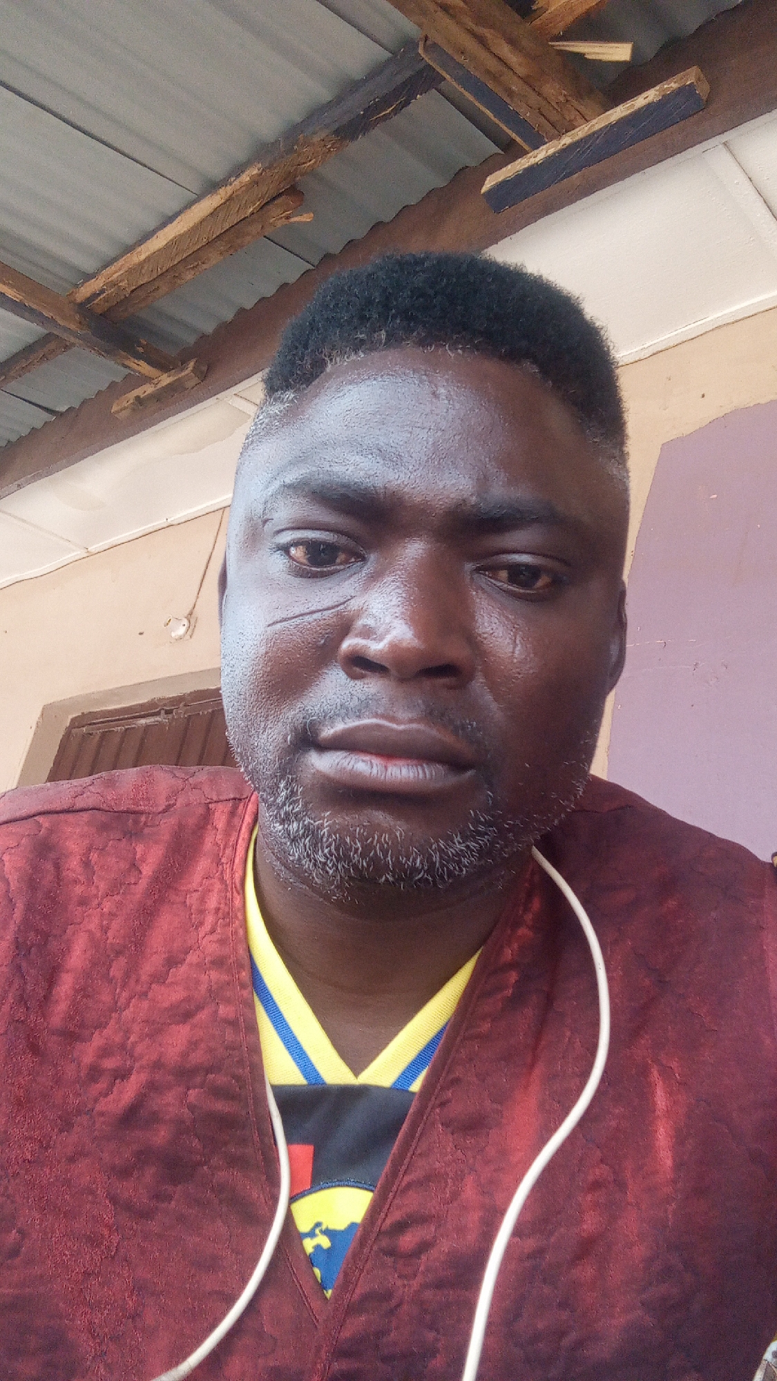 A close-up shot features a man with dark skin and a short, well-groomed haircut. A visible scar marks the left side of his face. He has a salt-and-pepper beard and a serious expression. He wears a maroon patterned top over a yellow and blue collared shirt, and a white earphone cord is visible around his neck. The setting appears to be indoors, with a corrugated metal ceiling and wooden beams suggesting a basic structure. Given the request for information about Sabon Tasha, Nigeria, the image likely depicts a person within this urban area, though specific details linking the image directly to a known location in Sabon Tasha are absent from the visual context alone.