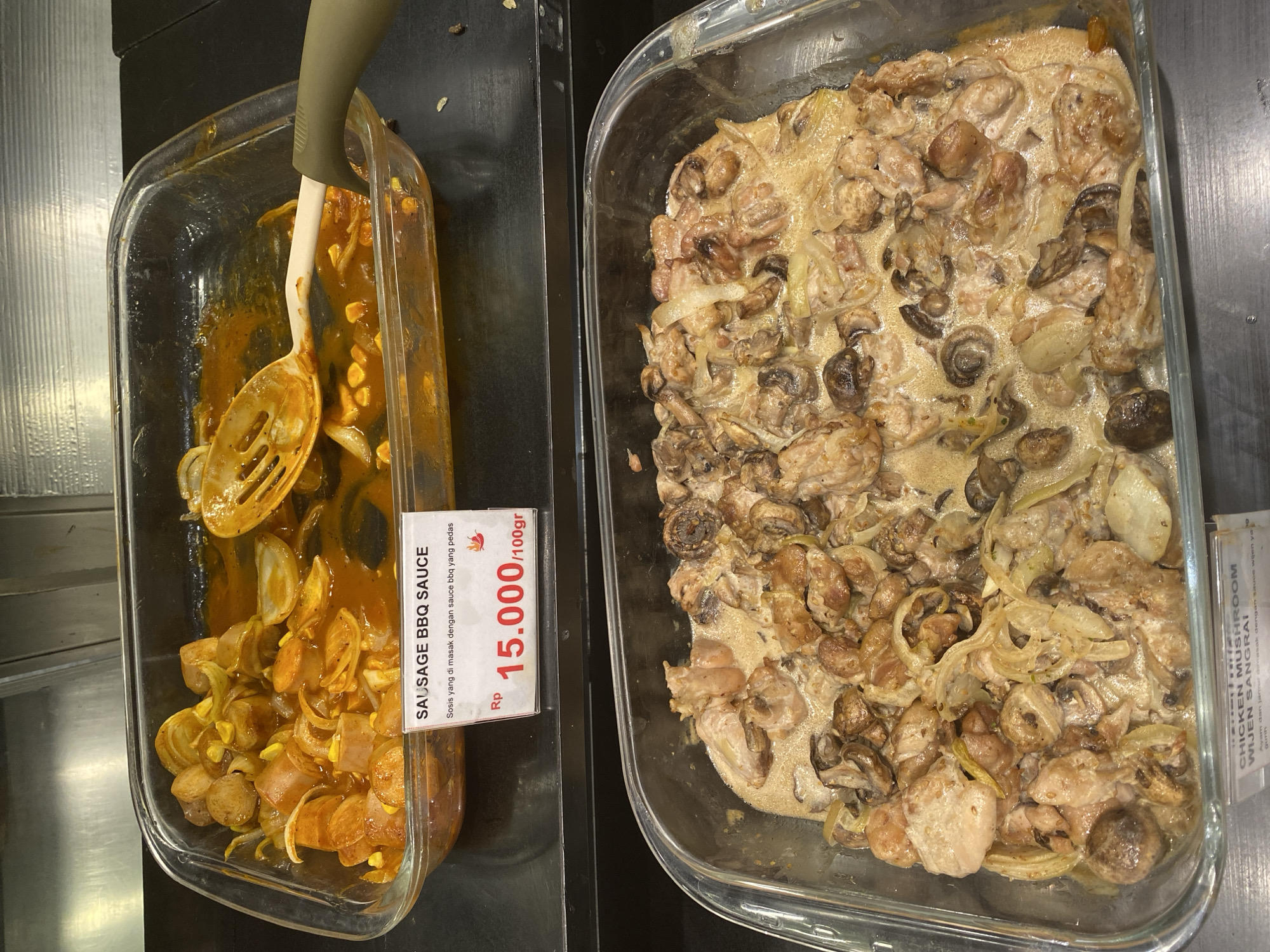 The image shows two glass rectangular dishes containing food, likely in a buffet or food service setting. The top dish contains sliced sausages and onions in a reddish-brown sauce, with a serving utensil in the dish. A white label in front of this dish reads "SAUSAGE BBQ SAUCE" and "Sosis yang di masak dengan sauce bbq yang pedas" which translates to "Sausages cooked with spicy BBQ sauce". Below this, it states the price as "Rp 15.000/100gr". The bottom dish contains chicken pieces and mushrooms in a creamy sauce with onions. A label in front of this dish reads "CHICKEN MUSHROOM WIJEN SANGRAI" and "Ayam dan jamur dimasak dengan sauce wijen ya gurih" which translates to "Chicken and mushrooms cooked with savory sesame sauce". The setting appears to be indoors, with a dark countertop and a metallic background, possibly a commercial kitchen or a restaurant. The lighting suggests overhead lights. The image was taken in East Jakarta, Indonesia.