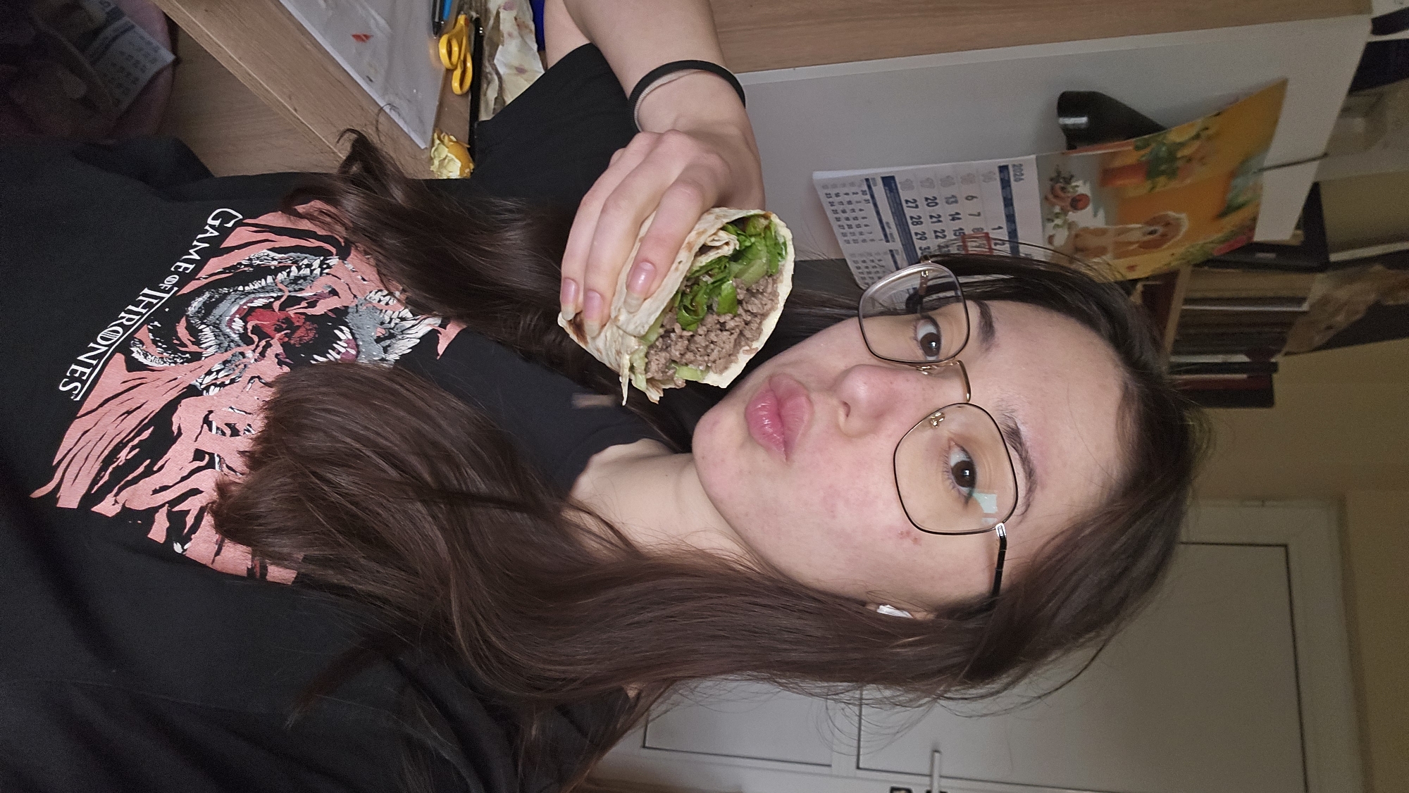 A person is holding a wrap in their right hand. The person is wearing glasses and a black shirt with a pink tiger on it. Part of the "GAME OF THRONES" logo is visible on the shirt. The person has long brown hair and is looking at the camera. A calendar is visible behind the person. The date "2026" is on the calendar. Part of the dates "6, 7, 8, 13, 14, 15, 20, 21, 22, 27, 28, 29, 30, 31" are visible on the calendar. A stapler and some papers are also visible behind the person. The person is in an indoor environment. Based on the visible items, it is not possible to determine the exact location in the city of Stara Zagora, Bulgaria.