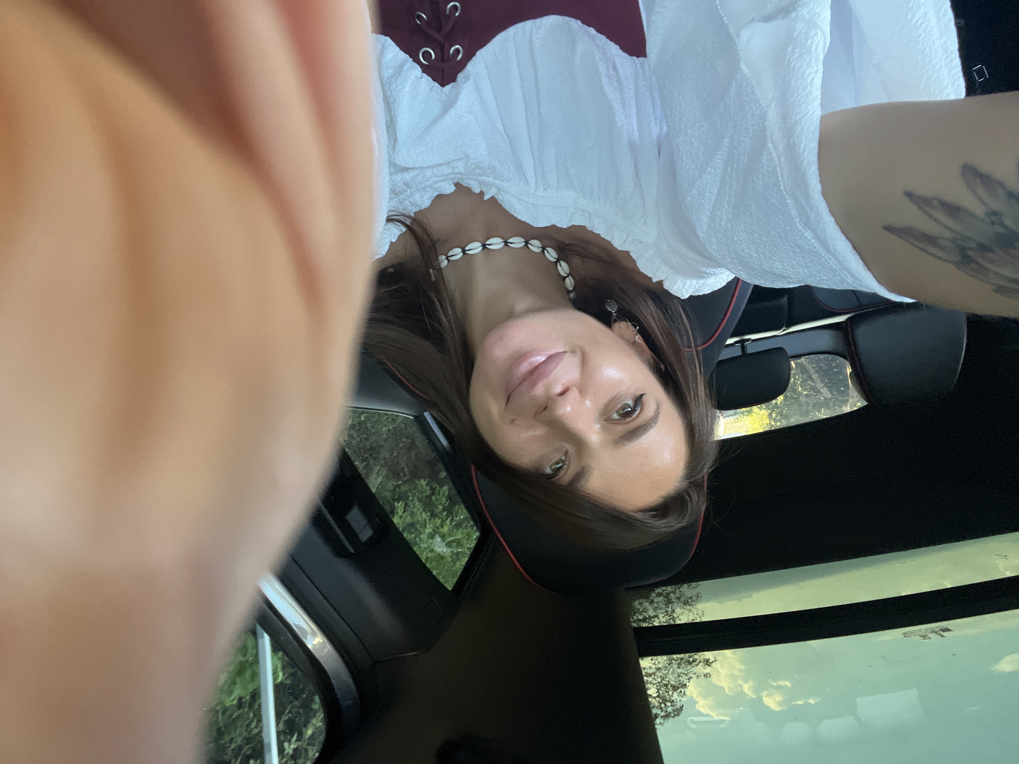 A woman is sitting in the driver's seat of a car. She is looking at the camera, and smiling. She is wearing a white shirt with a maroon corset over it, and a shell necklace. There is a tattoo on her left arm. The car has a sunroof, and the sky is visible through it. The car is parked in a wooded area.