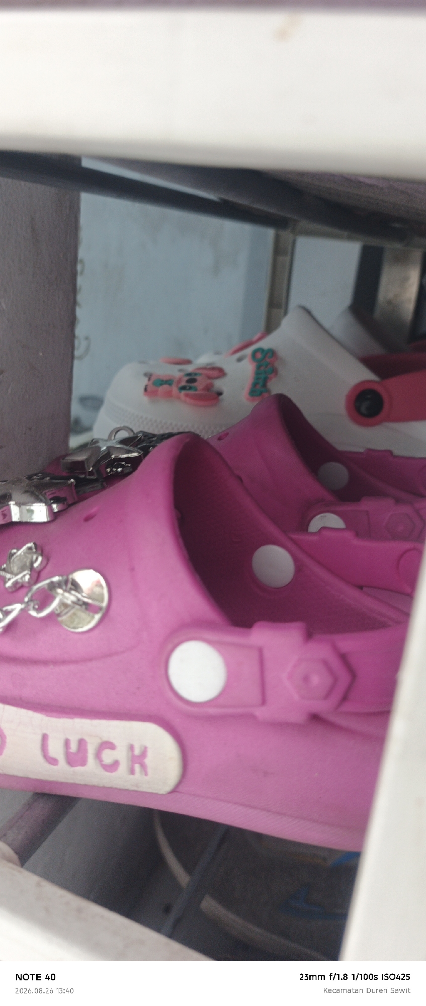 The image shows a close-up, angled view of two pairs of pink and white slip-on shoes, possibly Crocs, with decorative charms. The shoes are on a shelf or ledge, with the pink shoes in the foreground and the white ones behind. The pink shoes have white accents and a white inscription that appears to read "GOOD LUCK". Silver-colored charms, including a star and a chain link, are attached to the pink shoes. The white shoes have pink and green decorative elements, including what looks like a stylized rabbit or animal shape and the word "Swift". The background is out of focus and appears to be a wall or structure. Text at the bottom of the image indicates "NOTE 40", a date "2026.08.26 13:40", "23mm f/1.8 1/100s ISO425", and "Kecamatan Duren Sawit".