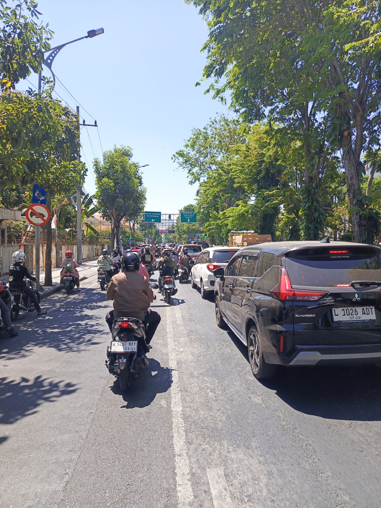 The image depicts a street scene in Surabaya, Indonesia, characterized by moderate traffic congestion on a sunny day. Several motorcycles and cars are visible on a multi-lane asphalt road lined with lush green trees. Pedestrians are not clearly visible, but motorcyclists, many wearing helmets, are the primary subjects in motion. One motorcycle in the foreground bears the license plate "W 5467 NAY G1-31". A black Mitsubishi Xpander SUV with license plate "L 1026 ADT" is in the right lane. Traffic signs, including a blue sign with a red circle and a diagonal bar indicating "No U-turn", and green directional signs are present. The sky is clear blue. The environment suggests an urban setting with ample foliage, indicating a well-maintained streetscape.