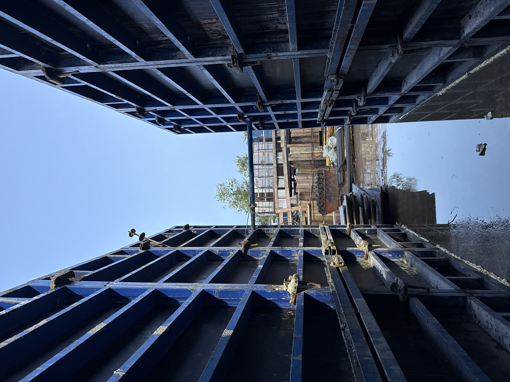 The image depicts a construction site in Moscow, Russia, with a narrow passageway formed by two tall, blue, modular formwork walls. The formwork is constructed from metal beams forming a grid pattern, with some visible signs of wear and tear, including rust and concrete residue. The ground between the walls is wet and reflective, indicating the presence of standing water, likely from recent rain or an ongoing construction process.
In the background, beyond the formwork, various construction materials and equipment are visible. These include stacks of rebar, piles of white bags (possibly cement or sand), wooden planks, and a large red dumpster. Further back, structures with scaffolding and what appears to be a building under construction can be seen. The sky above is clear and blue, suggesting fair weather. No people are visible in the frame. The overall scene conveys a sense of active construction work in an urban environment.