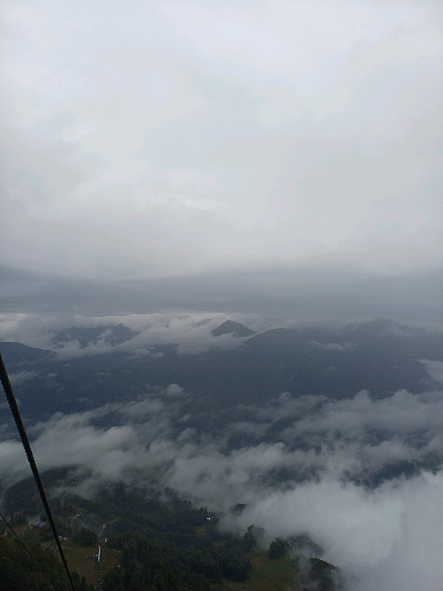 The image shows a mountainous landscape with thick clouds covering the peaks, suggesting a cloudy or foggy day. In the foreground, a cable for a ski lift or gondola is visible, angled diagonally from the upper left to the lower left of the frame. Below the cable, the slopes of the mountain are covered with dense green trees. There appear to be some structures or buildings scattered on the mountainside, possibly part of a ski resort or village. The lower portion of the image depicts a green, forested mountain slope with a winding path or road. The middle ground reveals layers of mountains partially obscured by clouds, creating a sense of depth and mystery. The distant background is dominated by more cloudy skies and the hazy silhouettes of mountain ranges. The scene captures a natural, rugged environment, likely at a high altitude, with evidence of human infrastructure like the ski lift cable.