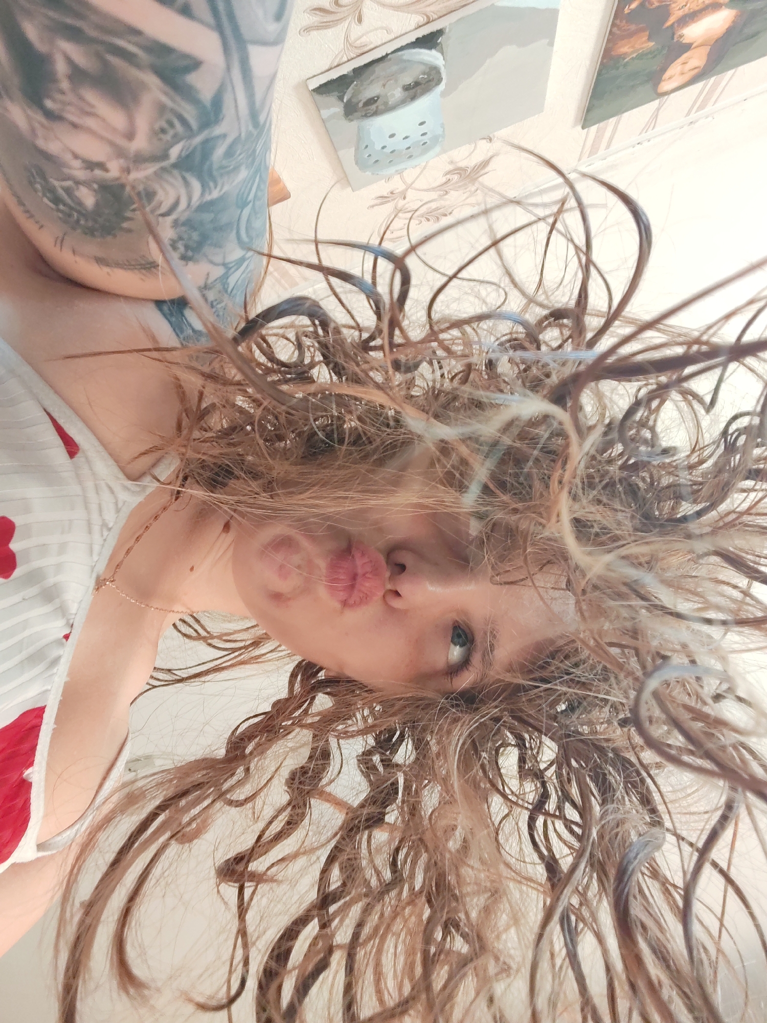 A low-angle, close-up photograph shows a person with long, curly, wet hair. The person's face is visible, exhibiting a puckered expression. A tattoo is visible on the upper left arm. A gold chain is around the neck. The person is wearing a white top with red heart-shaped designs. The background consists of a white wall with two framed pictures. The location and city are unknown.