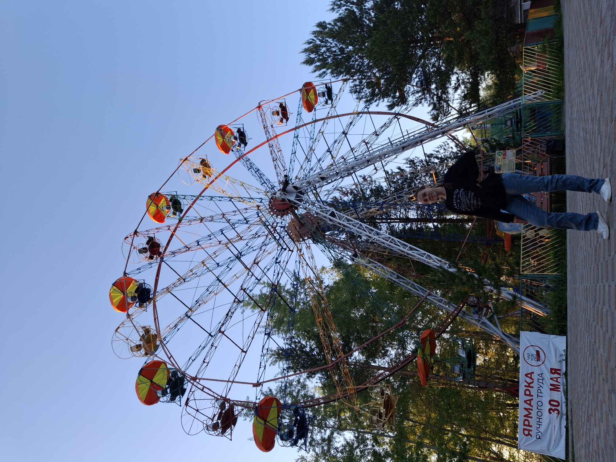 A Ferris wheel dominates the frame, set against a clear blue sky. The Ferris wheel has a complex network of red, yellow, blue, and white steel struts. Each passenger car has an umbrella top, colored red, yellow, and green. People are seated in the cars. A young woman stands in the foreground. She is wearing a black sweatshirt, blue jeans, and white sneakers. She is smiling and looking toward the camera. In the background, there are green trees. A sign in Russian with the words "ЯРМАРКА ручного труда 30 МАЯ" is located on the right side of the frame. The scene takes place in the city of Chaikovsky, Russia.