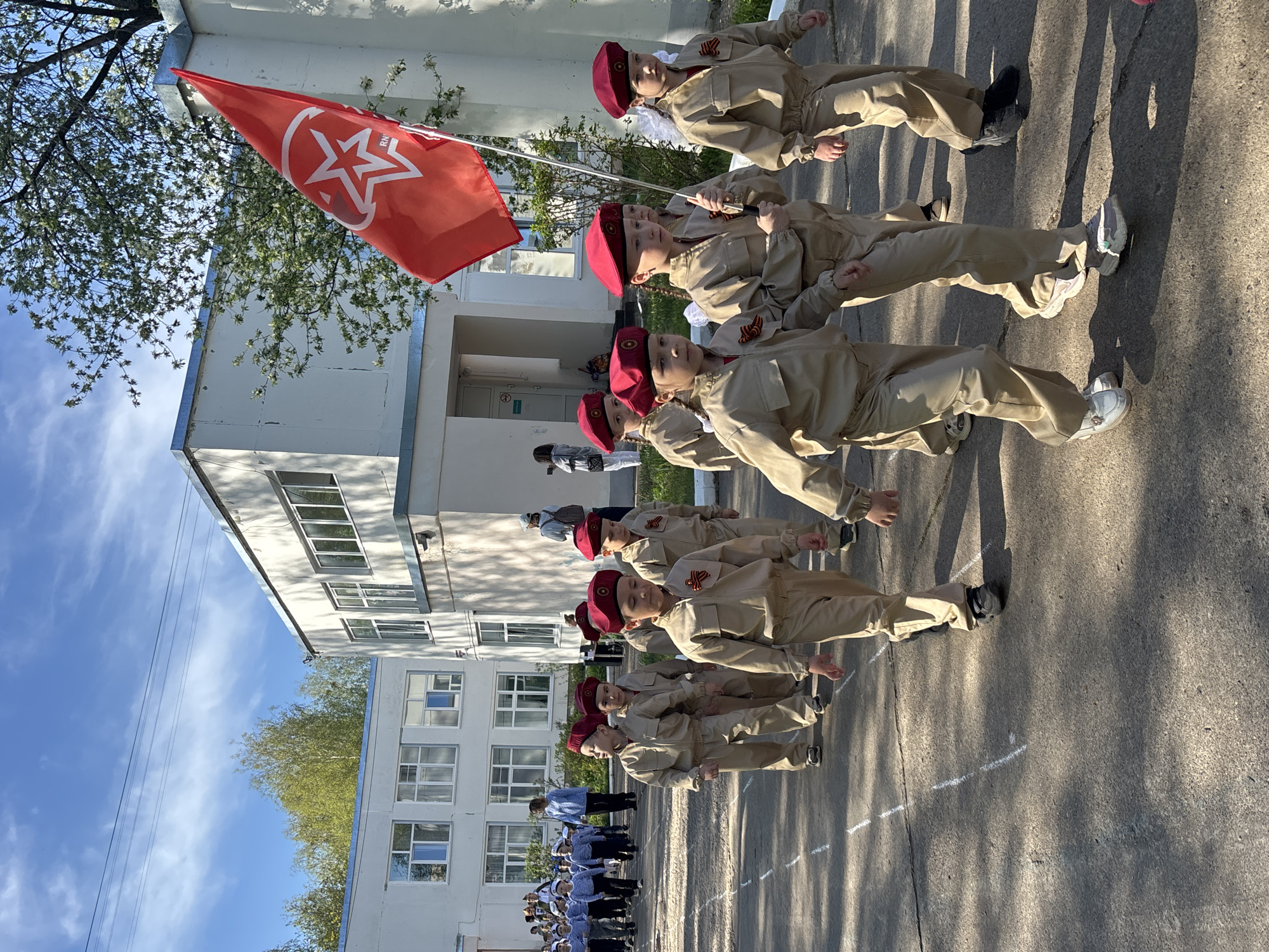A group of children dressed in uniforms and red berets stand in formation, holding a red flag with a white star and crescent moon on it. They are lined up on a concrete surface with white markings, possibly for alignment. Behind them is a multi-story building with numerous windows. In the distance, more people in striped shirts and white caps are visible. The sky is blue with some clouds, and trees are visible in the background.
The image was captured in the city of Chaikovsky, Russia. The attire and flag suggest some kind of historical or ideological event involving children, possibly related to youth organizations or commemorative activities.