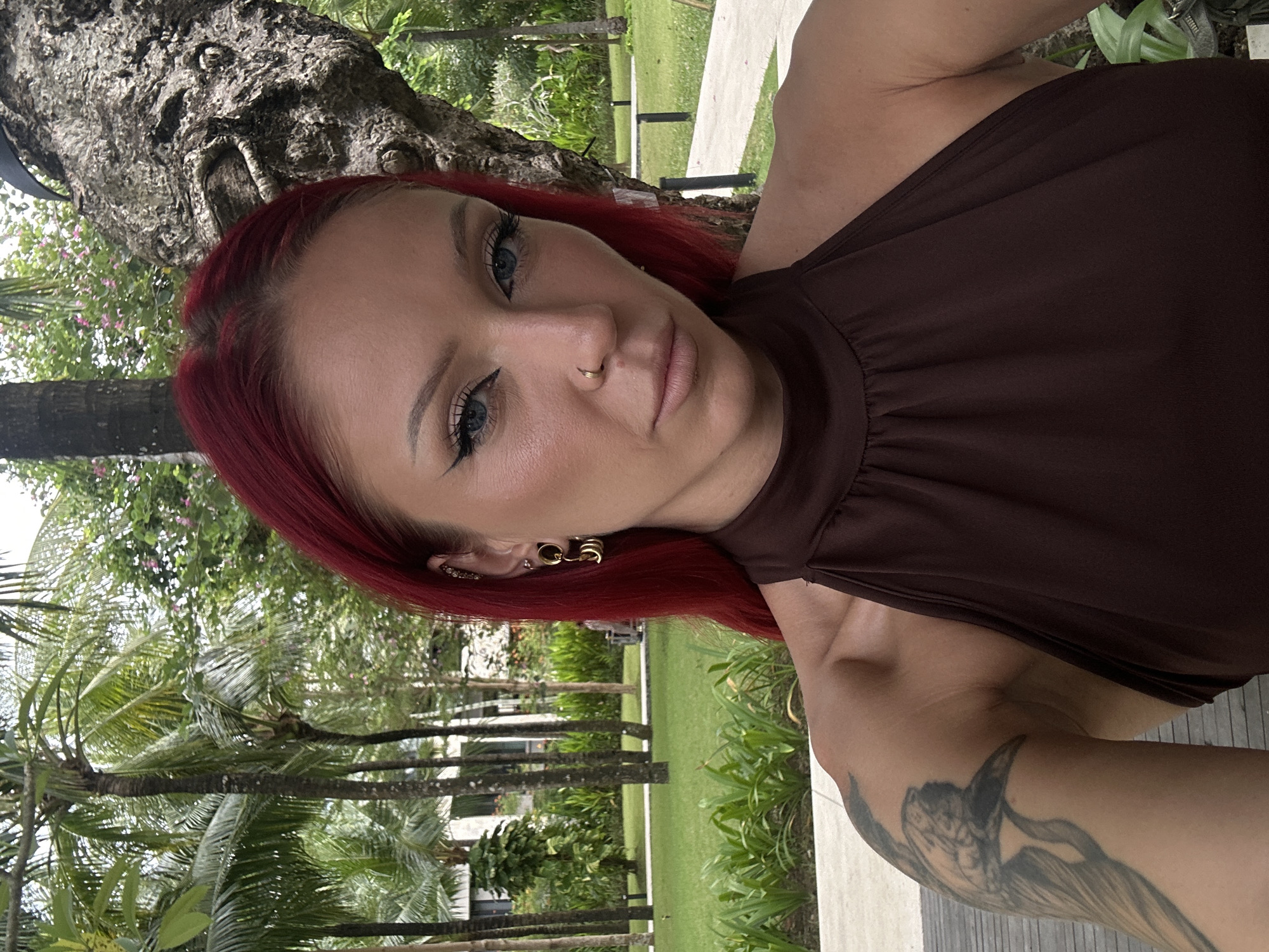 The image depicts a selfie taken by a fair-skinned woman with red hair, light blue eyes, and a nose piercing. She is wearing a brown halter top. Her left arm, featuring a visible tattoo, is extended to hold the camera. The background reveals a tropical environment suggestive of Nusa Dua, Indonesia. Palm trees and other lush greenery, along with glimpses of architecture, confirm the location's nature. The shot is composed at a slight angle.