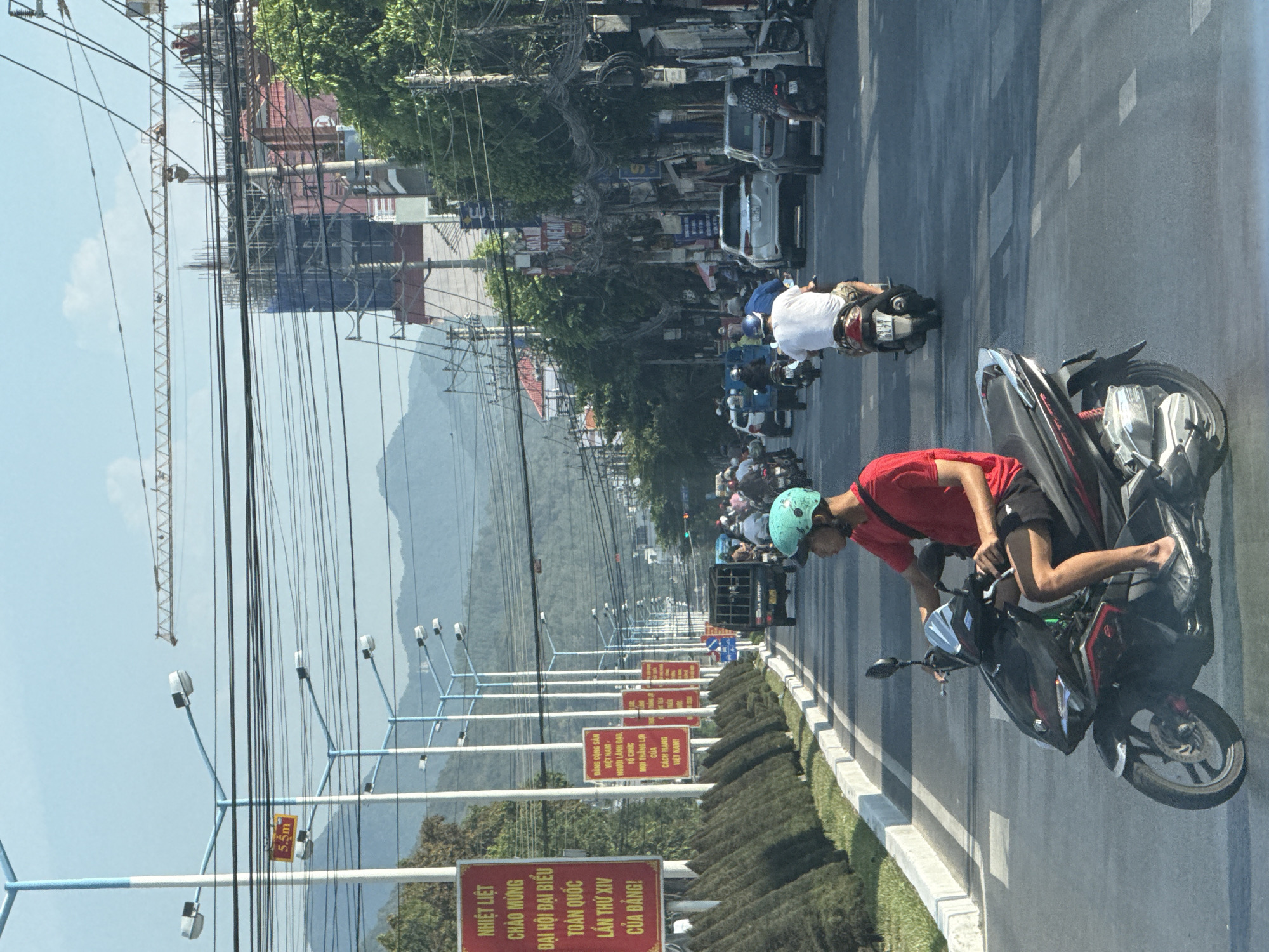 The image shows a street scene in Bac Nha Trang Ward, Vietnam. A man wearing a red shirt and a teal helmet is riding a black motorcycle in the foreground. A series of red signs with yellow lettering line the road, and streetlights are visible along the road. In the background, other motorcycles and vehicles are traveling along the street. Buildings and a crane are visible in the distance, along with mountains. Numerous power lines run across the scene.
