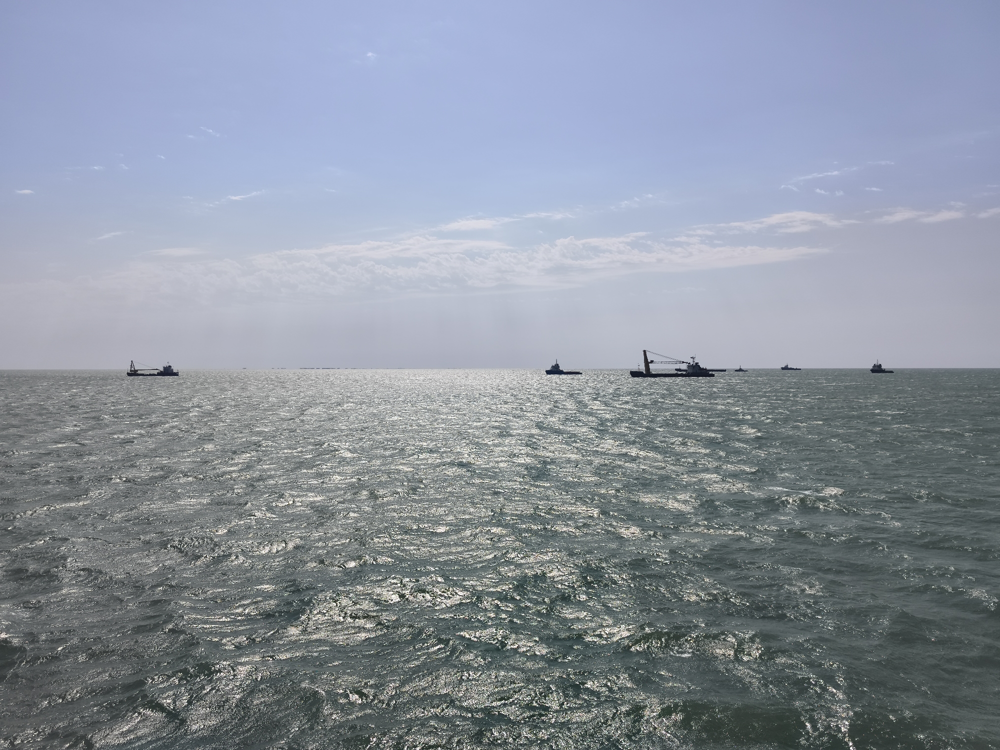 The image shows a wide expanse of choppy, dark green ocean under a pale blue sky with scattered white clouds. Several ships of various sizes are visible on the horizon, silhouetted against the sky. In the foreground, the water reflects the sunlight, creating a shimmering effect. The scene is calm and open, with no land in sight. The location is Pirallahi Raion, Azerbaijan.