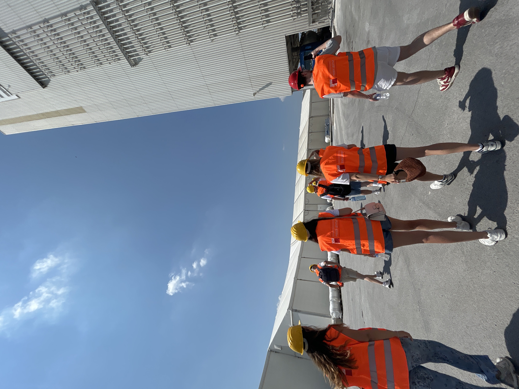 In Elin Pelin, Bulgaria, a group of six individuals, four women and two men, are walking on an asphalt surface under a clear blue sky with scattered clouds. They are wearing bright orange safety vests over their clothing and hard hats. The vests have reflective stripes and lettering in what appears to be a Cyrillic script, indicating they are likely part of a tour or work crew at an industrial or construction site.
To their left, a large white tent structure stretches into the distance, with large rolls of white material visible inside. To their right, a tall, modern building with a corrugated metal exterior and vertical piping details rises against the sky. The building has a clean, industrial aesthetic.
The group is walking away from the camera, with the majority facing towards the large building. One individual on the far right is wearing a red hard hat, distinguishing them from the others who are wearing yellow. One of the women in the foreground is carrying a brown woven bag, and another is holding a clear plastic bottle of water. The overall scene suggests an organized visit or educational tour of a facility in Elin Pelin.