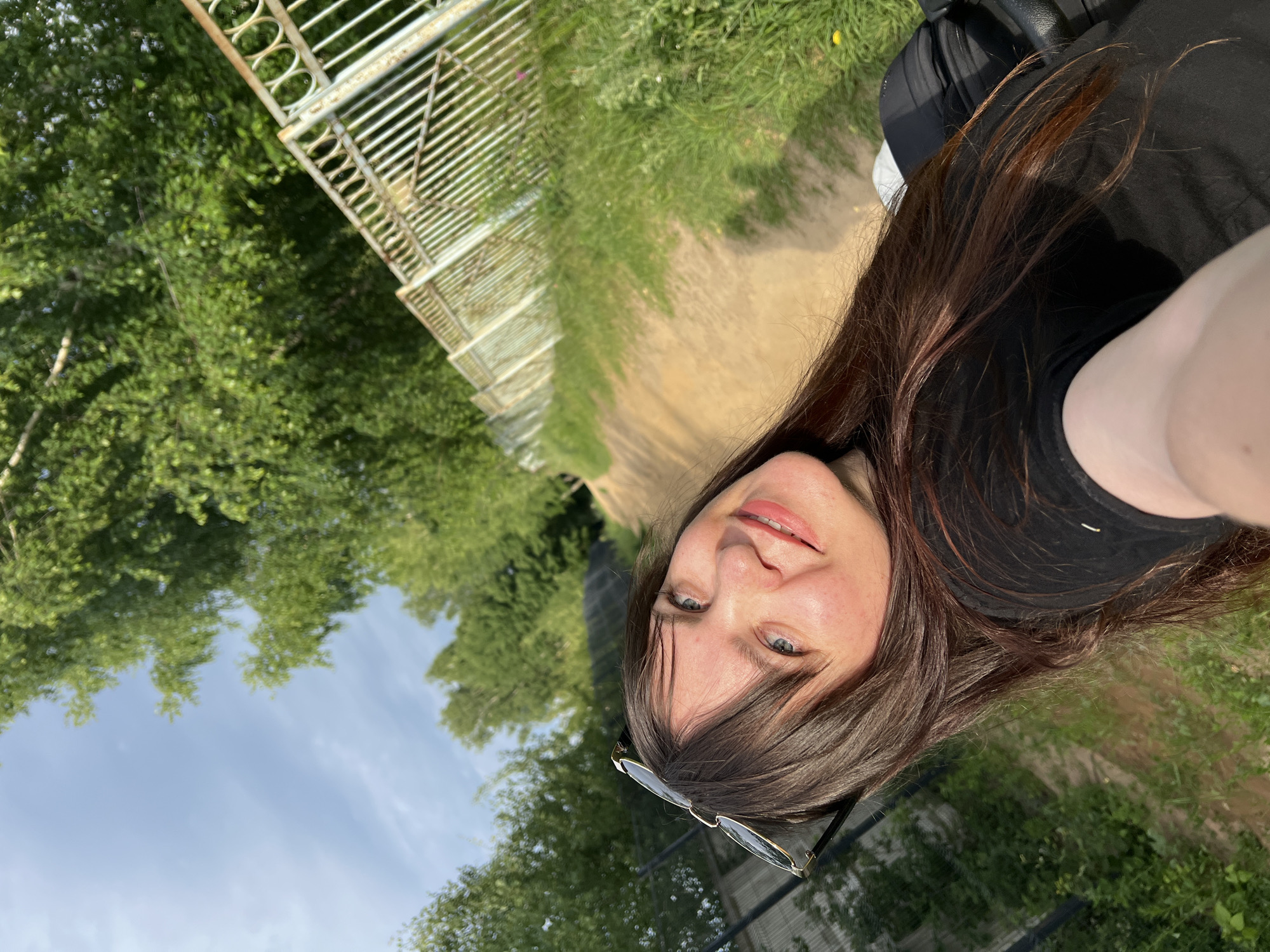 A woman with long brown hair and blue eyes is taking a selfie outdoors. She is wearing sunglasses on her head and a black t-shirt. She is smiling and looking at the camera. The background shows a dirt path with a white metal fence on the right and lush green trees on the left. The sky is blue with some clouds. The image appears to be taken in a park or rural area. The location is identified as Noviy, Russia.