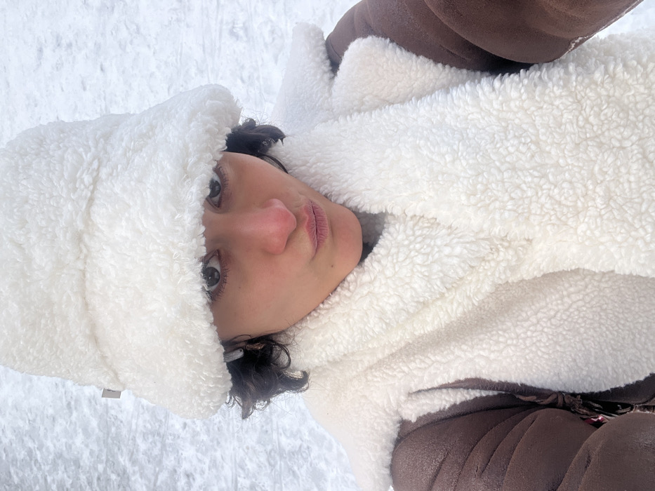 A person is depicted in a close-up selfie, facing directly towards the camera, against a textured white background consistent with snow or ice. The individual has dark, curly hair visible beneath a white, fluffy, sherpa-style bucket hat. Their eyes are dark brown, and their lips are pursed in a slight pout. The nose appears reddish, indicative of cold exposure. They are wearing a white, fluffy, sherpa-style scarf, which is wrapped around the neck and partially covers a brown jacket or coat with a visible sherpa lining or trim. A light-colored earbud is present in the left ear. The scene takes place outdoors in cold, snowy conditions in Paris, France.