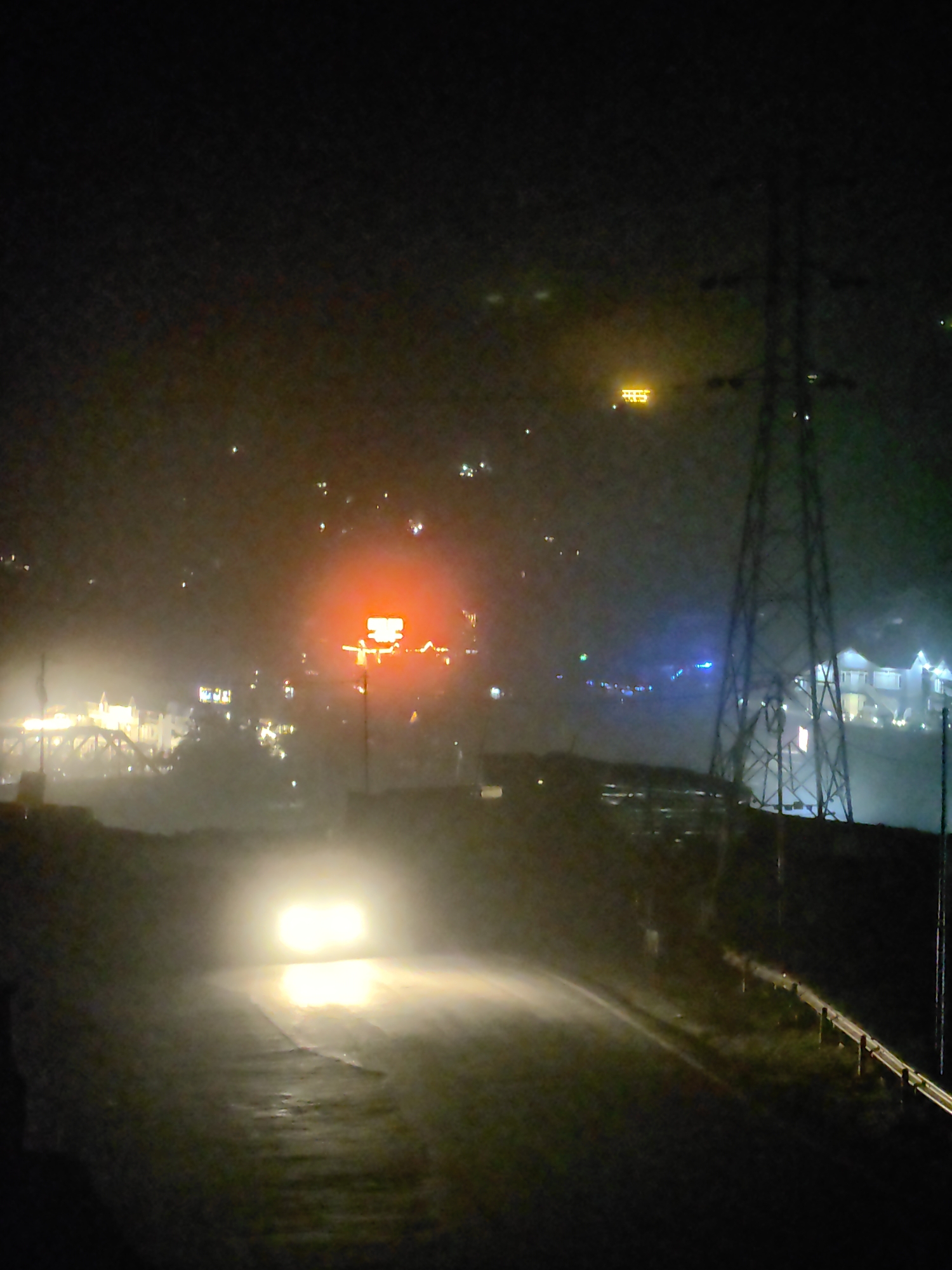 This is a night-time photograph taken on a foggy, possibly wet, road in Kulang, India. A car with its headlights on is approaching the viewer on the road, casting a bright glow. In the background, there are various lights from buildings and structures, including a prominent, bright orange illuminated sign that appears to be a billboard or advertisement. A large electricity pylon is visible to the right of the frame. The overall atmosphere is dark and somewhat obscured by mist or fog.