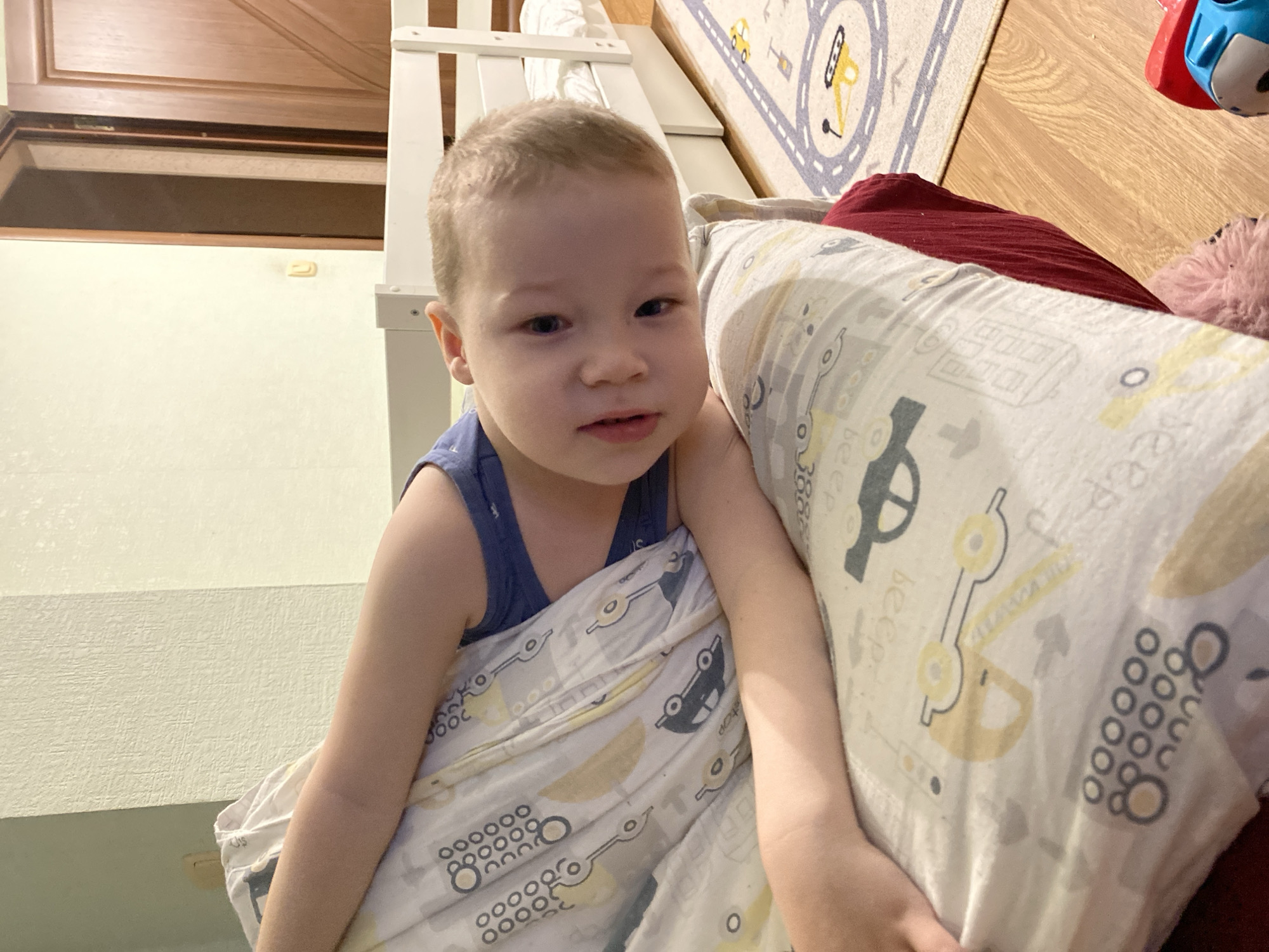 A fair-skinned toddler is sitting propped up by a large pillow. The child wears a dark blue tank top. The pillow has a white background and is printed with drawings of construction vehicles. Above and behind the child, a light-colored wooden structure is partially visible. Behind this structure, a door is seen. A child's rug with a circular design sits on a wooden floor, a blue and red toy is also partially visible. The image shows the child in an indoor setting. There is no indication of a specific location in Sochi, Russia.