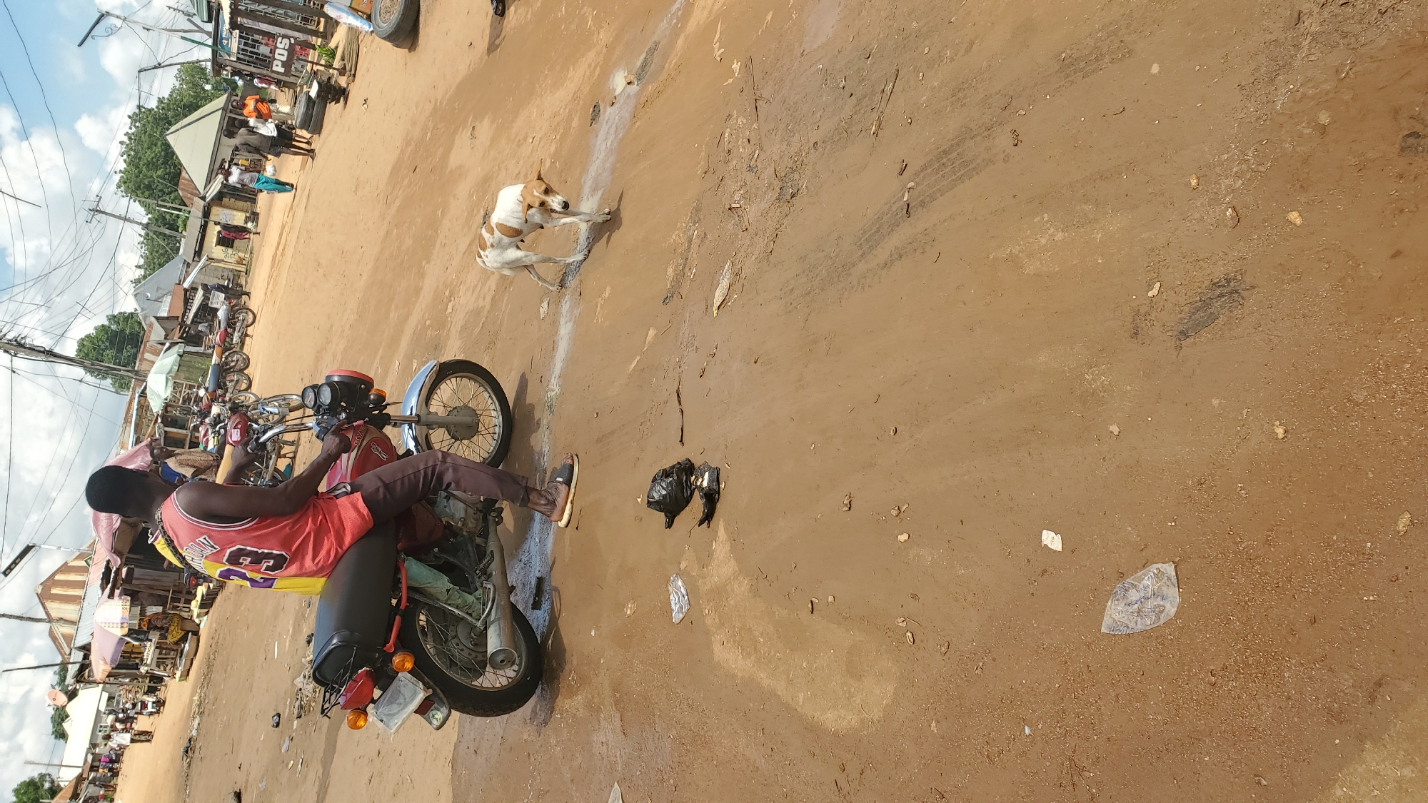 A man is sitting on a red motorcycle in Jalingo, Nigeria. He is wearing a red basketball jersey with the number 23 on it and dark pants. The motorcycle is parked on a dirt road. A small, white and brown dog is walking in front of the motorcycle. In the background, there are several market stalls, more motorcycles, and people walking. The sky is cloudy. There is some litter on the ground, including a black plastic bag.