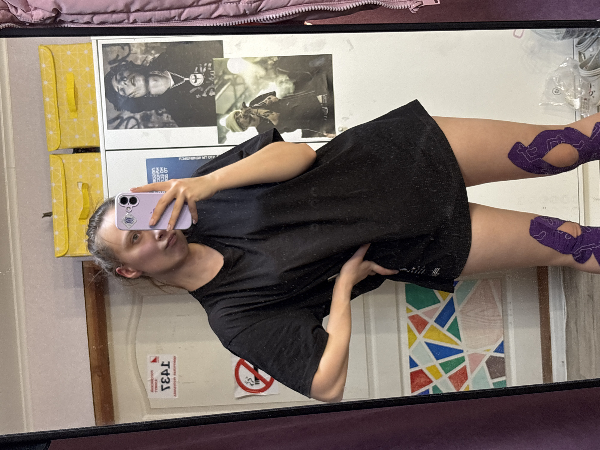 A young woman captures a mirror selfie indoors, appearing to be in a personal room or dwelling. She holds a light purple smartphone with a sticker on the back, and her face, partially obscured by the phone, shows a neutral to slightly pouting expression. She is wearing a black t-shirt or dress. Notably, her upper thighs and hips are adorned with purple kinesiology tape featuring a repetitive stylized pattern.
The background, reflected in the mirror, reveals several details. To the left, two yellow storage bins with white patterns are stacked. Above them, two framed black and white posters are visible; the top one depicts a person with long hair and a cross pendant. Below these, a white wall or door features a colorful geometric design reminiscent of a mosaic or stained glass. A red circular "no smoking" sign with a crossed-out cigarette is affixed to the wall. Partially visible text on a blue object behind the phone reads "ЛО ЦВ НПШ" and "КОМНАТА" (ROOM), with other indistinct mirrored characters. Below the "no smoking" sign, mirrored text "Т4Э1" and smaller words, possibly "ТОЛЬКО" (ONLY) and "МОСКВА", are visible on a white surface. Part of a pink quilted jacket is visible at the top edge of the frame, and white shoes in plastic packaging are in the upper right corner. The lighting suggests an indoor setting with no specific time of day discernible. While the location is given as Прикамский, Russia, the visual cues, such as the Russian text, align with this context.
