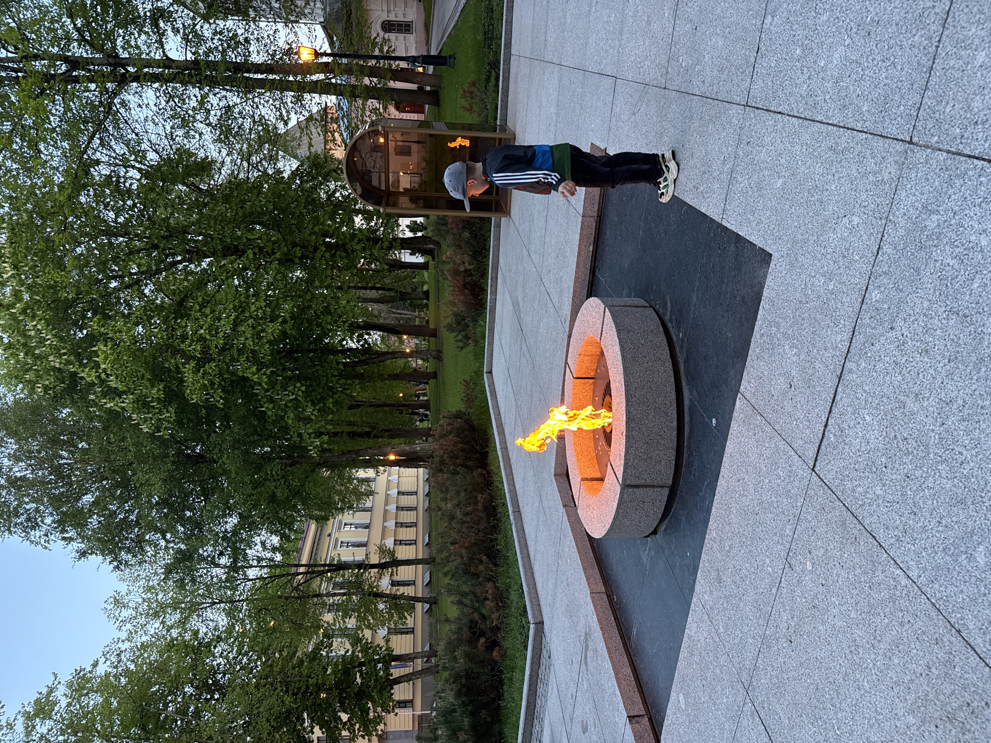 The image captures a scene in Veliky Novgorod, Russia. A young boy wearing a baseball cap, a blue and black jacket with white stripes, dark pants, and athletic shoes stands near the Eternal Flame memorial. The Eternal Flame burns within a circular stone structure set into a dark, granite-paved area. Behind the boy is a small metal-and-glass shelter also containing a flame. The area around the memorial is landscaped with trees and grass, and a multi-story building is visible in the background. The ground is paved with gray stone. It appears to be dusk or early evening.