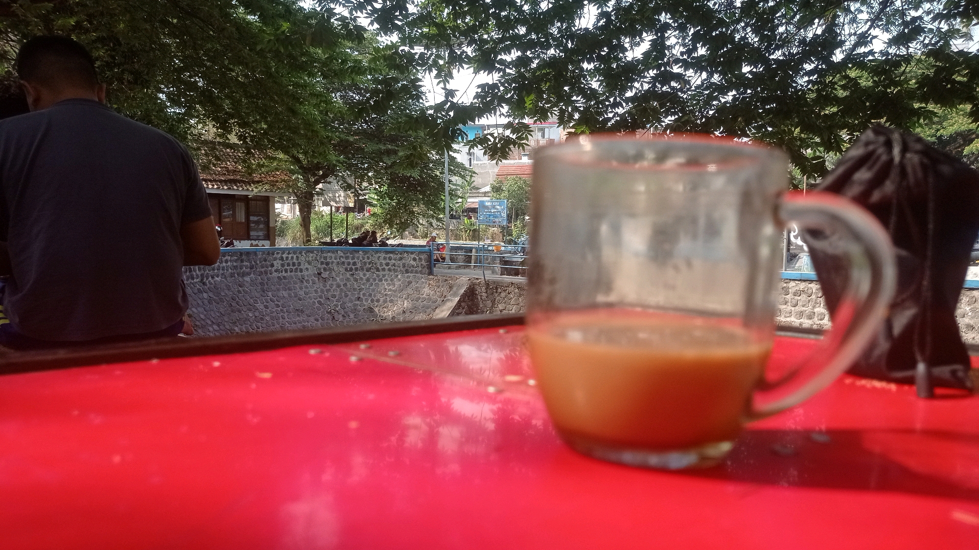 The image depicts an outdoor scene at the Mulyoagung, Indonesia. In the foreground, a clear glass mug filled with a light brown liquid, likely coffee or tea, sits on a vibrant red surface. To the left, a person is seated with their back to the camera, wearing a dark t-shirt and shorts. Behind them, a stone wall borders a body of water or canal. In the background, lush green trees with dense foliage create a natural canopy, with a glimpse of buildings and a blue sign visible through the leaves. A black bag is positioned to the right of the mug. The lighting suggests it is daytime, with sunlight filtering through the trees.