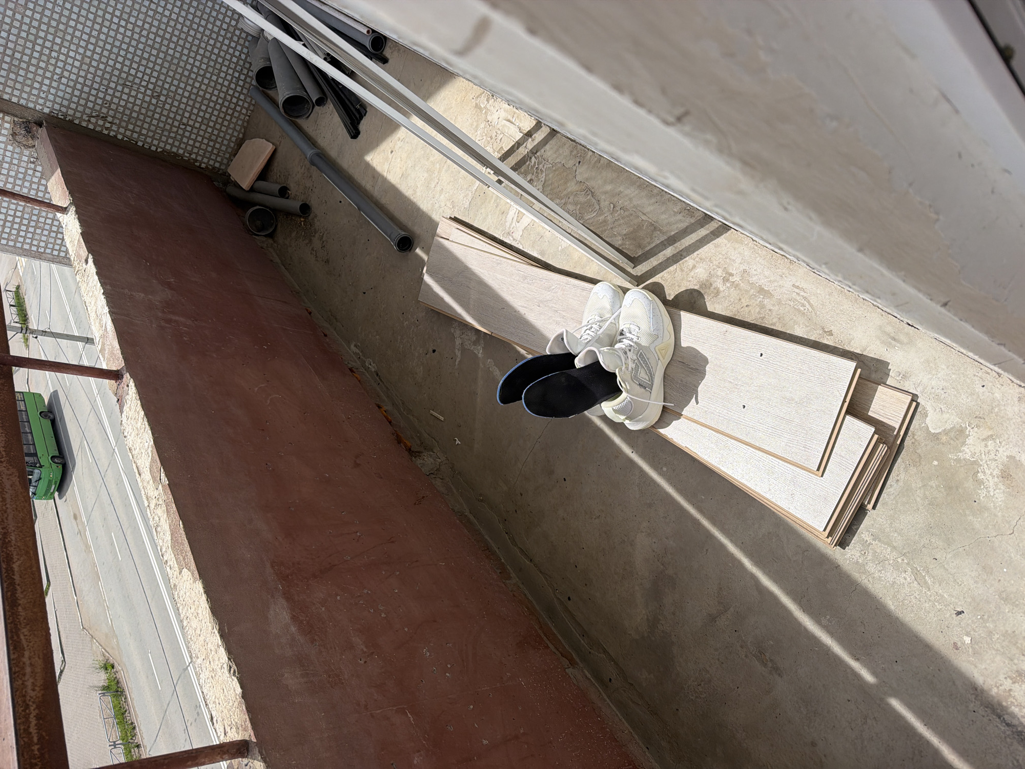 This high-angle shot looks down from a balcony or loggia in an urban or suburban environment, likely in Chaikovsky, Russia. The concrete floor of the balcony is cluttered with various items. A pair of light-colored (white or very light grey) sneakers with black insoles rests on a stack of light-toned wooden planks, consistent with laminate flooring. Nearby, several grey pipes of different diameters are visible, along with some white metal bars, possibly part of a clothes drying rack or construction materials. A small reddish-brown tile also lies on the concrete. One corner of the balcony features a section of small grey square floor tiles.
To the left, a weathered reddish-brown wall or railing forms the edge of the balcony, offering a view down onto a two-lane road. A green bus is visible driving on this road, and a short metal fence lines the roadside. The presence of building materials on the balcony suggests ongoing renovation or storage.
Strong, distinct shadows are cast across the concrete floor, indicating bright, sunny weather and a time of day when the sun is relatively low, likely morning or late afternoon. No people are visible in the image, and there is no discernible text. The scene captures an ordinary moment, providing a factual glimpse into the immediate surroundings and the street below.