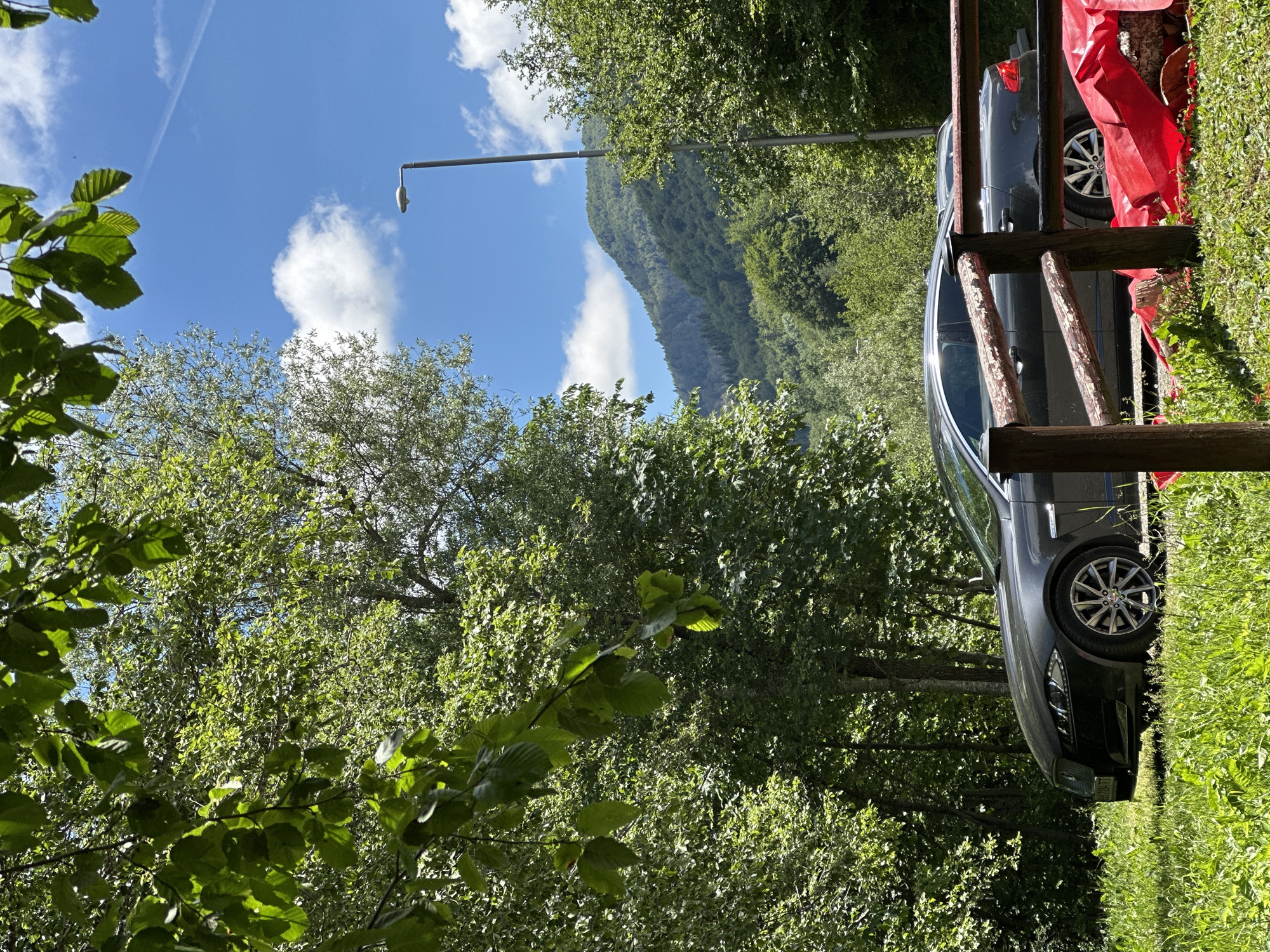 An eye-level shot captures a dark gray Jaguar sedan parked on a grassy area beside a rustic wooden fence. The car is angled towards the right, with its front left wheel and part of the driver's side visible. Behind the car, a dense forest of green trees stretches out, with a prominent, tall streetlamp positioned to the right of the vehicle. In the background, a majestic, tree-covered mountain rises under a clear blue sky dotted with fluffy white clouds. A thin white contrail is visible in the upper right portion of the sky. The scene is bathed in bright sunlight, casting shadows and highlighting the vibrant green of the foliage. The location is identified as Bare Kraljske, Montenegro.