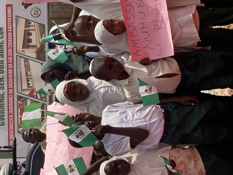 This image depicts an outdoor gathering in Ifira, Nigeria. The scene is dominated by a dense group of approximately 15-20 individuals, primarily children and young adolescents of African descent, standing in bright daylight.
**People:** The majority are children, estimated to be between 6 and 14 years old. Many wear what appears to be a school uniform, consisting of white collared shirts (some long-sleeved, some short) and dark green trousers or skirts. Several girls wear white hijabs or other head coverings, with one girl wearing a dark green hijab. Their expressions are generally neutral or attentive, with some looking directly at the viewer. An adult male, wearing a light-colored shirt, is partially visible in the upper left background, behind the children.
**Objects:** Numerous small Nigerian flags (green-white-green vertical stripes) on sticks are held aloft by the children. Additionally, several small green and white flags featuring the Nigerian Coat of Arms are visible. One child on the right holds a pink handwritten sign with black text, partially legible as "AMBASSAD NUHU BA LI BLO." In the background, a large banner displays the text "GWAMNA, SEN. UBA SANI, CON" and "ON INGATANCCEN JAGORANCI DA SADUAKAR DA KAI DON CIGABAN KAUYUKANMU" (Hausa for "On strong leadership and sacrifice for the development of our communities"). This banner also features architectural renderings of modern buildings and the seal of the Kaduna State Government. A second, partially obscured pink banner with "UBA SAN" in black letters is visible behind some children. A dark vehicle is partially visible on the far left.
**Actions/Interactions:** The individuals are standing in a crowd, holding up flags and signs, indicating participation in a public event or demonstration. Many appear to be posing or observing an unseen point in front of them.
**Environment:** The setting is outdoors, under clear, bright lighting conditions, suggesting sunshine. The ground surface is light-colored and appears to be dirt or gravel.