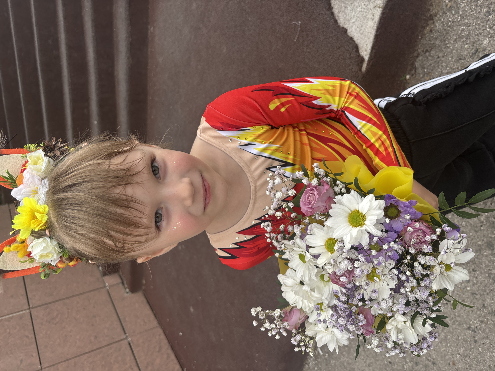 A young girl, approximately 5-8 years old, stands outdoors in Perm, Russia, looking directly at the camera with a subtle smile. She wears a long-sleeved gymnastics or dance leotard with a nude-colored torso transitioning into vibrant red, orange, and yellow flame-like patterns on the sleeves and sides, embellished with small rhinestones on the chest. Her head is adorned with a decorative headband featuring felt fox ears and a floral arrangement of yellow, white, and orange artificial flowers, some greenery, and small pinecones. Fine glitter is visible on her upper cheeks, below her eyes. She holds a large, mixed bouquet of flowers with both hands, positioned in front of her lower torso. The bouquet contains white daisies with yellow centers, light purple filler flowers, white baby's breath, and several light pink/lavender roses, wrapped in bright yellow paper at the base. She is also wearing black athletic pants with white stripes and ruffled detailing along the seams. The background consists of dark brown, horizontally grooved steps to her right and a reddish-brown textured wall to her left, suggesting an urban outdoor setting.