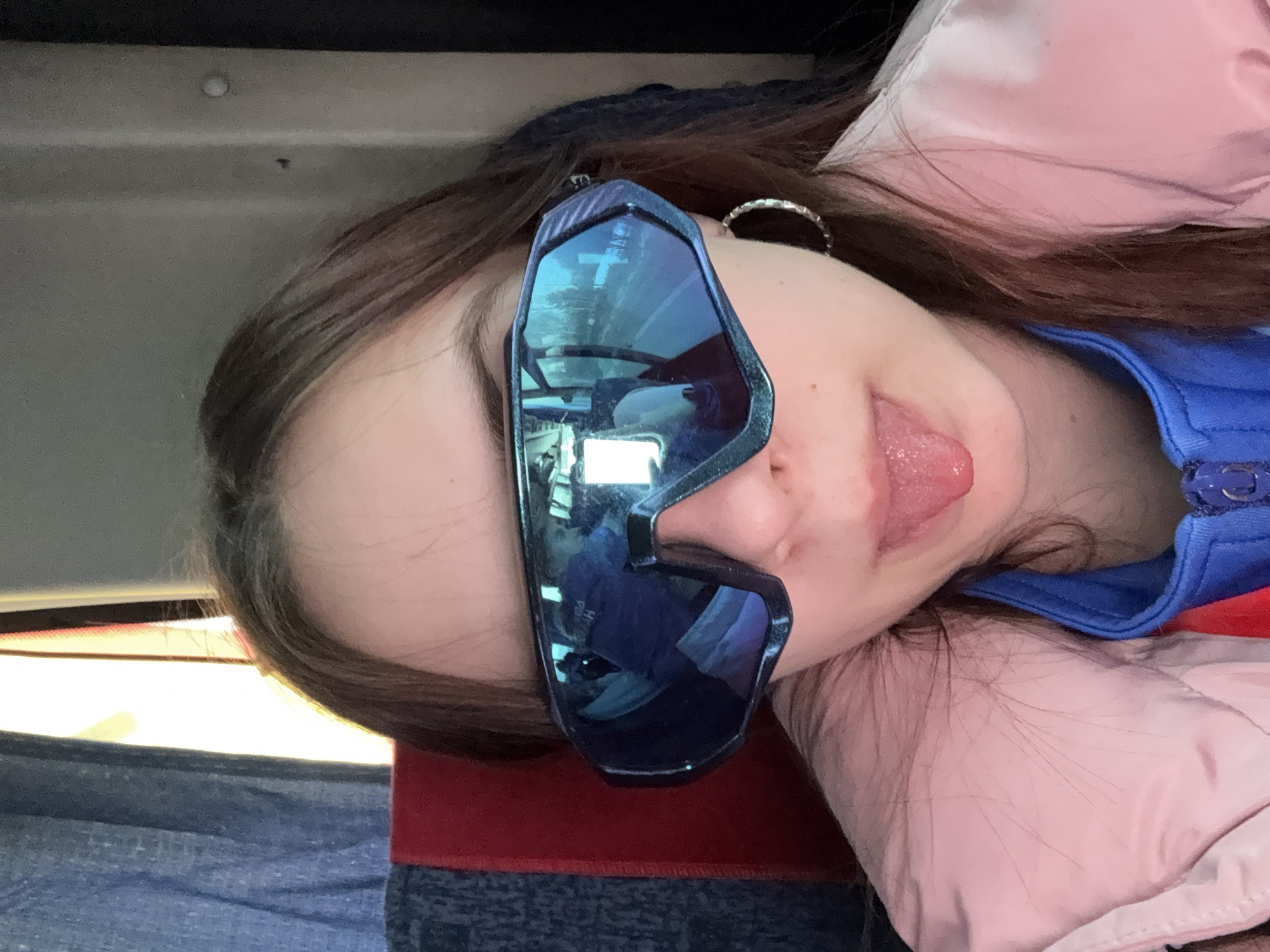 A young person lies sideways in what appears to be a vehicle, possibly a bus or train, based on the interior paneling visible. The individual is wearing reflective blue sports sunglasses and sticking their tongue out. They have brown hair and are wearing a pink jacket over a blue shirt. A metal hoop earring is visible. The location is identified as Челва, Russia; however, this identification appears to be inferred from external data, as there are no explicit geographical markers in the image itself.