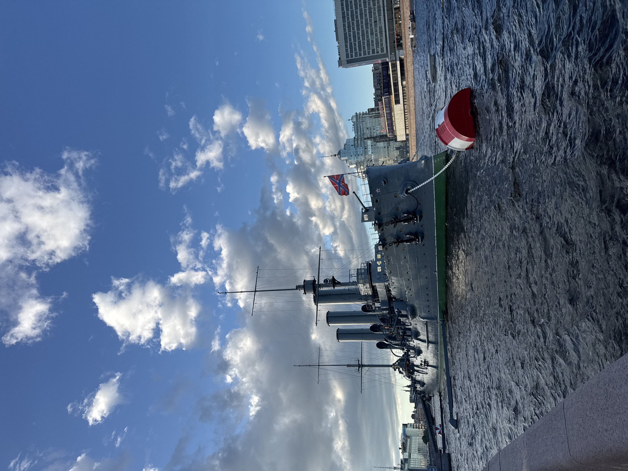 An image shows a wide shot of the Aurora cruiser docked on the Neva River in Saint Petersburg, Russia. The cruiser is grey with a green waterline and has three smokestacks. The Russian flag is flying from the stern. A red and white buoy is tied to the ship with a chain. The river is choppy, and the sky is blue with white clouds. In the background, modern buildings are visible along the riverbank, including the new facade of the historic Aurora. The foreground shows a stone embankment.