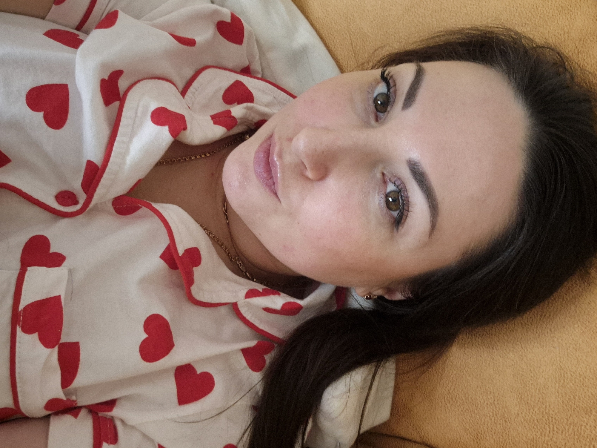 The image captures a woman lying down, looking directly at the camera with a soft, engaging gaze. Her head is tilted slightly, and her dark hair is spread out around her. She has brown eyes, well-defined eyebrows, and visible eyelashes, suggesting light makeup. Her lips are slightly parted, appearing glossy.
She is wearing white pajamas adorned with numerous red heart patterns of varying sizes. The garment features red piping along its edges, framing the collar and front opening. A thin, gold-colored chain is visible around her neck, resting on her skin beneath the pajama fabric.
The woman is positioned against a textured, light yellow-orange surface, which appears to be upholstery from a couch or bed. The lighting in the scene is soft and even, highlighting her features and the details of her clothing. The overall mood conveyed is relaxed and intimate, suggesting a casual moment indoors. There is no visible text in the image.