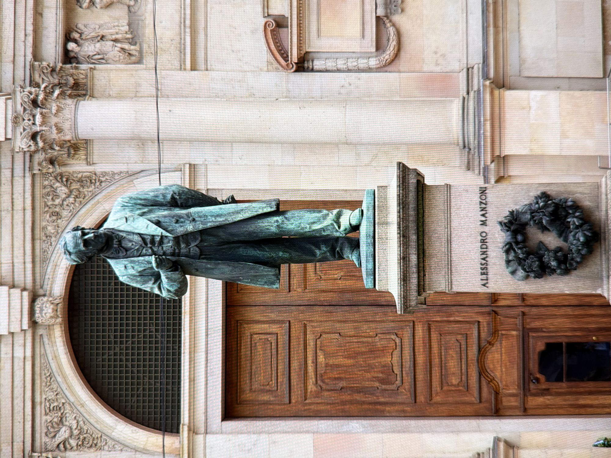 A full-body bronze statue of a man stands on a stone pedestal in front of a large wooden double door and a building facade. The statue depicts a man in a suit with a tie, his left hand in his pocket and his right hand extended slightly forward. The building facade is made of stone and features architectural details such as arches, columns, and decorative carvings. Below the statue, on the pedestal, the name "ALESSANDRO MANZONI" is inscribed, and a bronze wreath is affixed. The scene appears to be an outdoor monument.