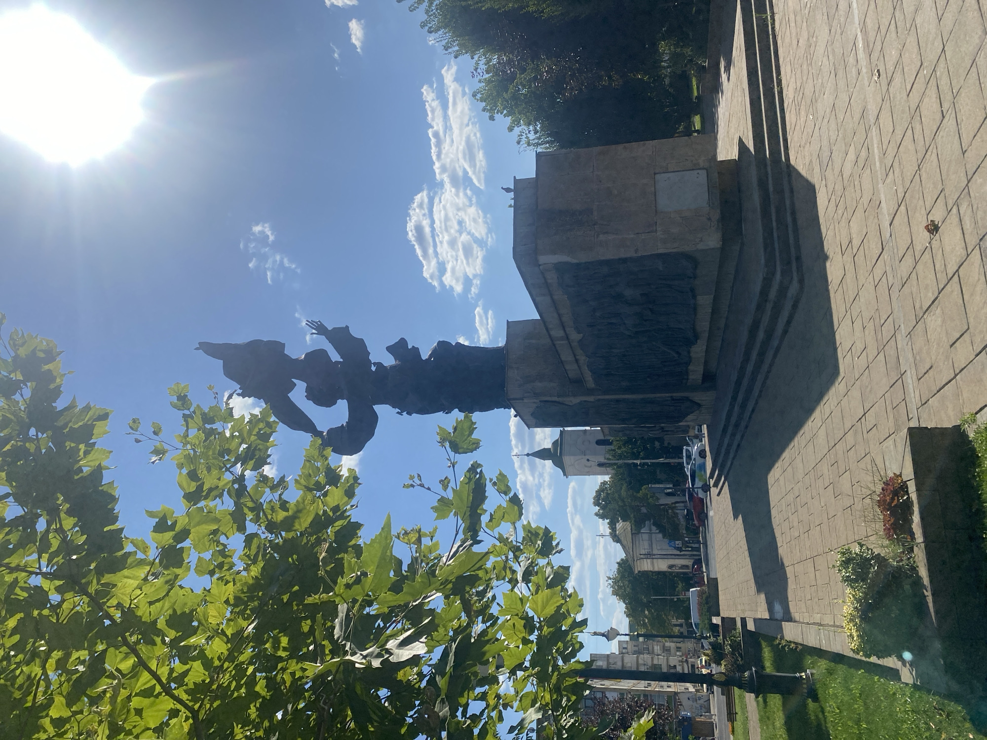 The image shows the "Monument of the Romanian Airmen" in Iași, Romania, located in front of the "National Theatre Vasile Alecsandri". The monument is a large, dark bronze sculpture depicting figures in flight and triumph. It stands on a stone pedestal adorned with bas-reliefs. In the foreground, a paved walkway with a pattern of stone tiles leads towards the monument. To the left, a lush green tree with large leaves partially obscures the view of the sky and the monument. The sky is a bright blue with scattered white clouds and a strong sun glare from the upper right corner. In the background, several buildings are visible, including a church with a tall spire and modern apartment buildings. There are no people visible in the immediate vicinity of the monument, though cars are seen on a street in the distance. The scene is bathed in bright sunlight, casting long shadows across the walkway.