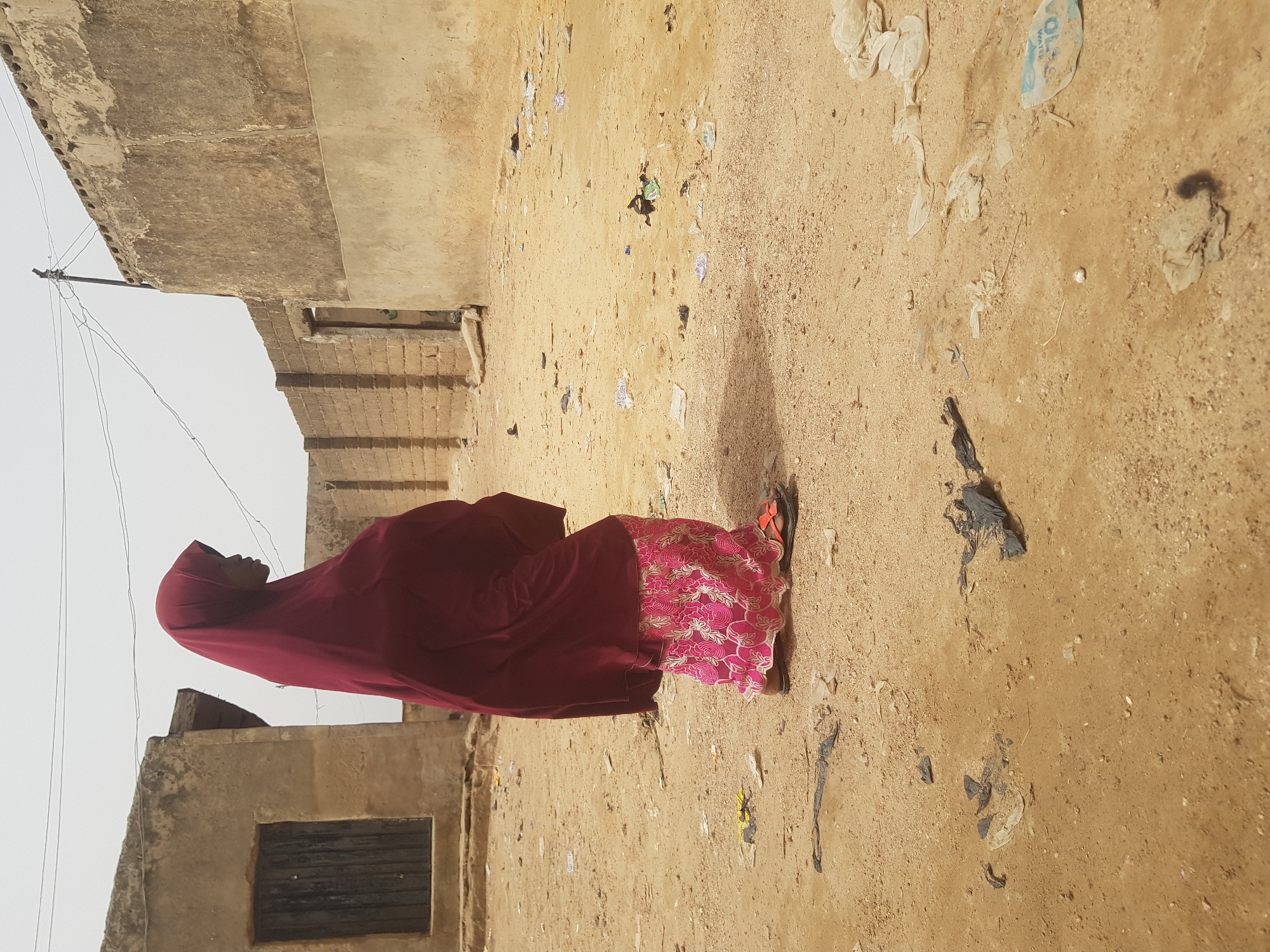 A woman wearing a maroon hijab and a pink patterned skirt stands on a dirt ground strewn with trash and debris. She is positioned between two aged, adjacent buildings with crumbling exteriors. The scene is set outdoors, likely within the urban environment of Dutsinma B, Nigeria.