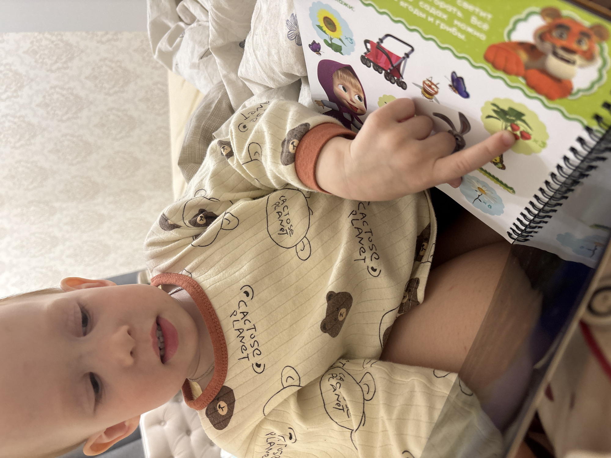 A young child is seated and looking down at a book. The child is wearing a tan long-sleeved shirt with a brown-trimmed collar and bear designs with the words "CACTUSC PLANET." The book is open to a page with illustrations including Masha from Masha and the Bear, a tiger, and various fruits and vegetables. The child's finger is pointing to a strawberry illustration in the book. The background is blurred, suggesting the scene is indoors, with some fabric or bedding visible in the periphery. The location is Ferma, Russia.