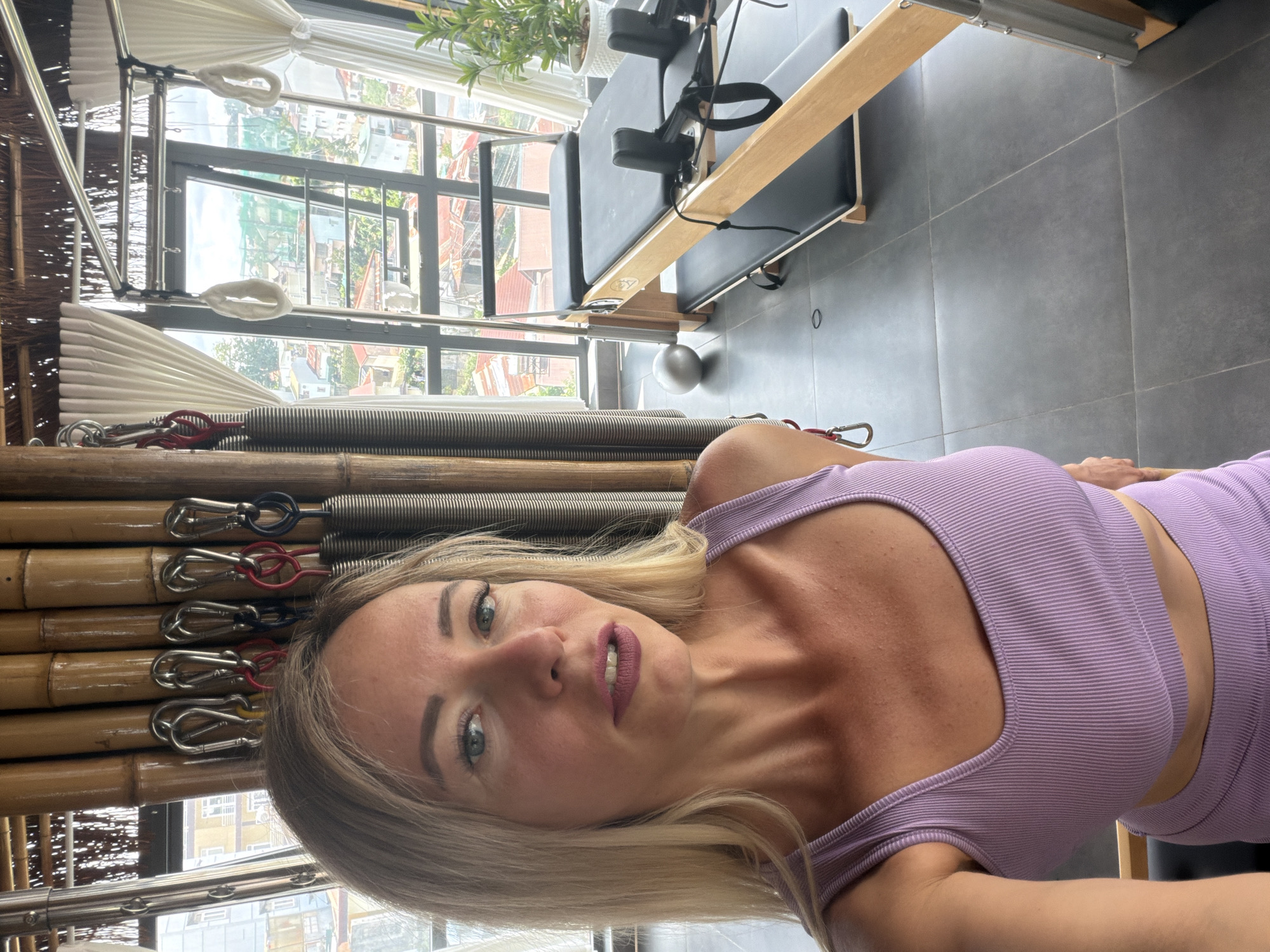 A blonde woman with blue eyes and purple lipstick takes a selfie in a room with a Pilates reformer. The room has a bamboo wall and large windows overlooking a city with red-tiled roofs, consistent with the architecture found in Da Lat, Vietnam. The Pilates reformer, a wooden apparatus with black padding and springs, is positioned by the window. A small silver ball and a potted plant are also visible in the room. The woman is wearing a purple ribbed crop top and skirt.
