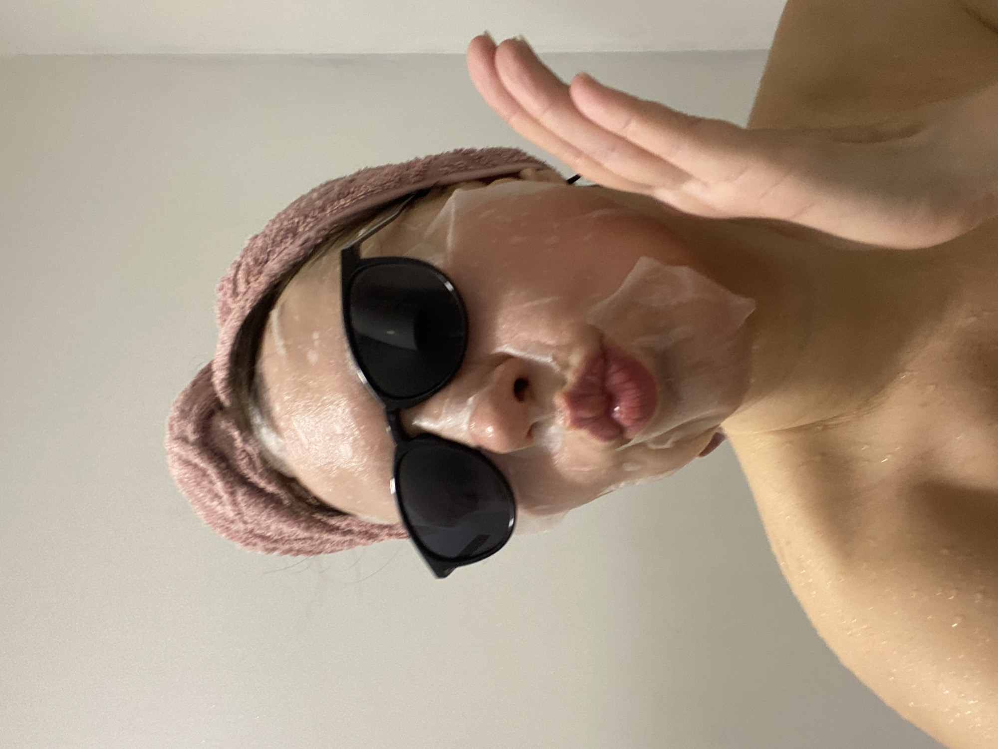 A woman is shown from a low angle, wearing a face mask and dark sunglasses, with a peach-colored towel wrapped around her head. Her skin appears wet, possibly from a recent shower. She is holding up one hand, with fingers extended and slightly curved. The background is a plain, light-colored wall. The location in Yekaterinburg, Russia is not evident from the provided image.