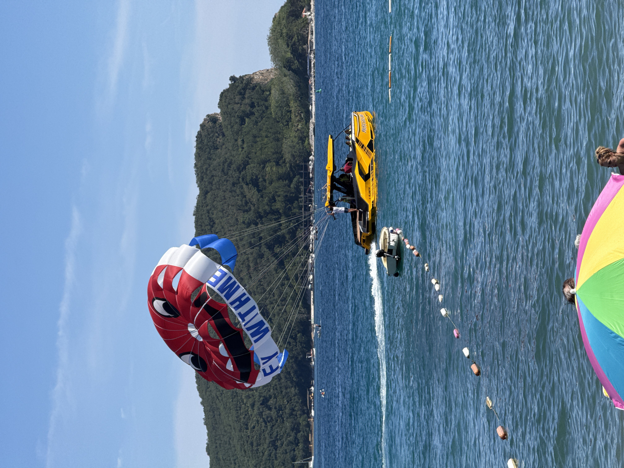 In the coastal city of Budva, Montenegro, a bright yellow parasailing boat is actively engaged in towing a brightly colored parachute over the clear blue Adriatic Sea. The parachute, featuring a playful design with a smiling face and the text "FLY WITH ME," is aloft in the sky, with the rope connecting it to the boat clearly visible. Several people are on the boat, observing the parasailer, while another small dinghy is moored nearby. In the foreground, a vibrant, multi-colored beach umbrella partially shields beachgoers from the sun, hinting at a popular tourist destination. The background reveals a lush, green, tree-covered hill, characteristic of the Budva coastline, with several other boats scattered across the water. The sky is a clear blue with a few wispy clouds, indicating fair weather.