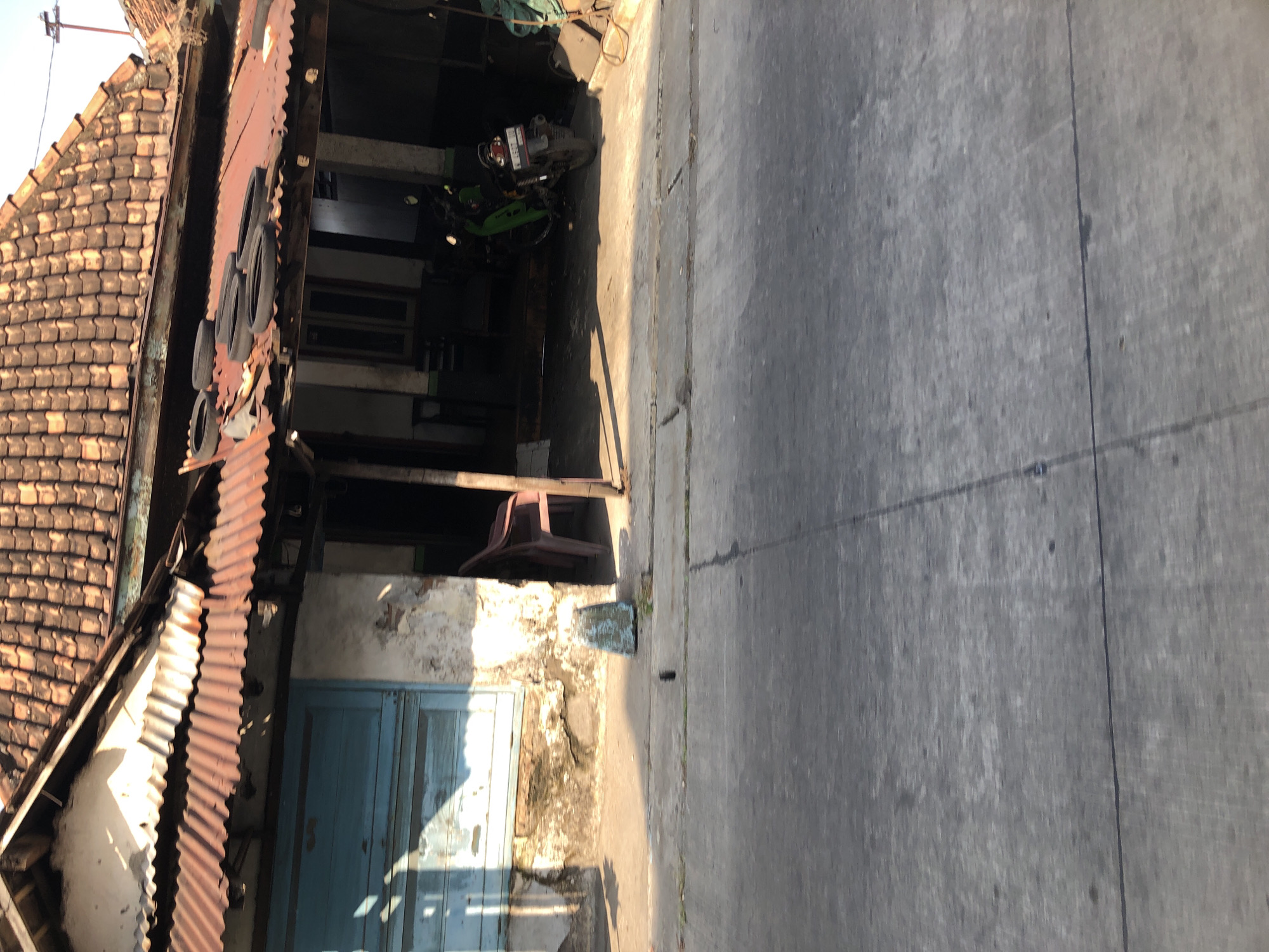 The image shows a weathered building with a corrugated metal roof and a tiled roof above. A blue wooden door is visible on the left side of the building. In front of the building, there is a concrete pathway. To the right, a green motorcycle is parked under an overhang. A plastic chair is positioned near the entrance of the building. The scene appears to be located in Ngaban, Indonesia, as indicated by the presence of what looks like typical local architecture and a motorcycle commonly seen in the region. The environment suggests an outdoor setting, possibly a street or alleyway, with the ground paved with concrete. The overall impression is one of a somewhat aged and utilitarian structure, possibly a shop or residence.