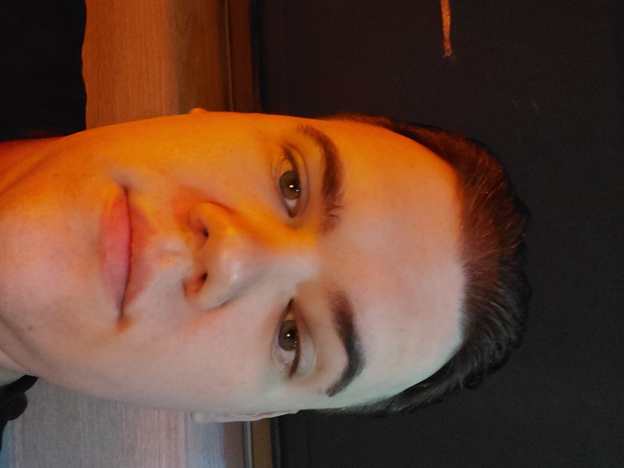 A close-up, indoor portrait of a person with fair skin, brown hair, and brown eyes is shown. The lighting is predominantly orange. The individual has full lips, a medium-sized nose, and well-defined eyebrows. The background is dark, but a wooden-textured surface is partially visible. There is no information available from the image that indicates a location in Tashkent, Uzbekistan. The person's actions and interactions are not evident from this image.