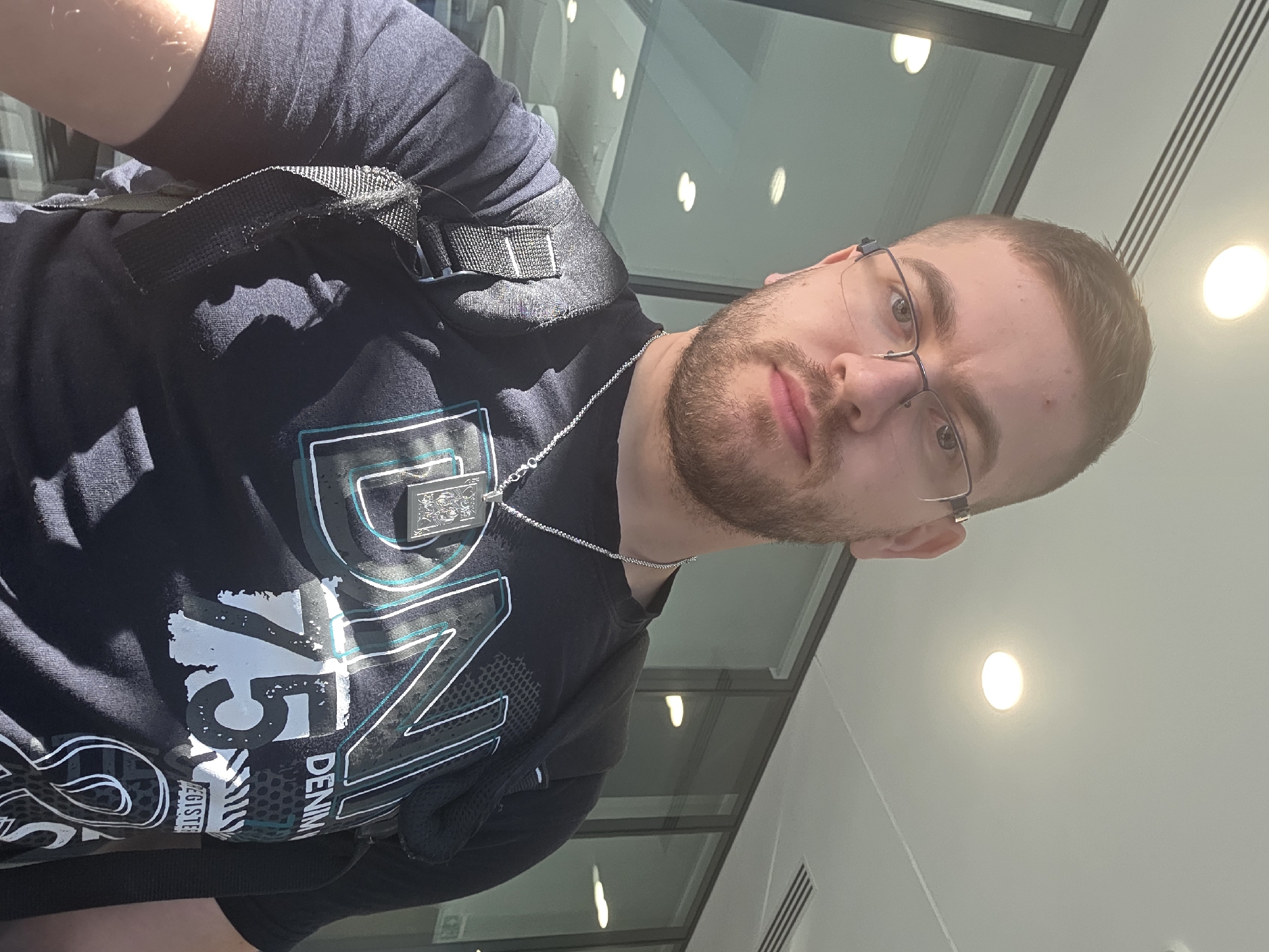 A man with short brown hair and a beard, wearing glasses, a dark t-shirt with a large design on the front, and a black strap across his chest, is facing the camera. He is indoors, under overhead lighting, with windows visible in the background. The location is unidentified and not confirmed to be in Plovdiv, Bulgaria.