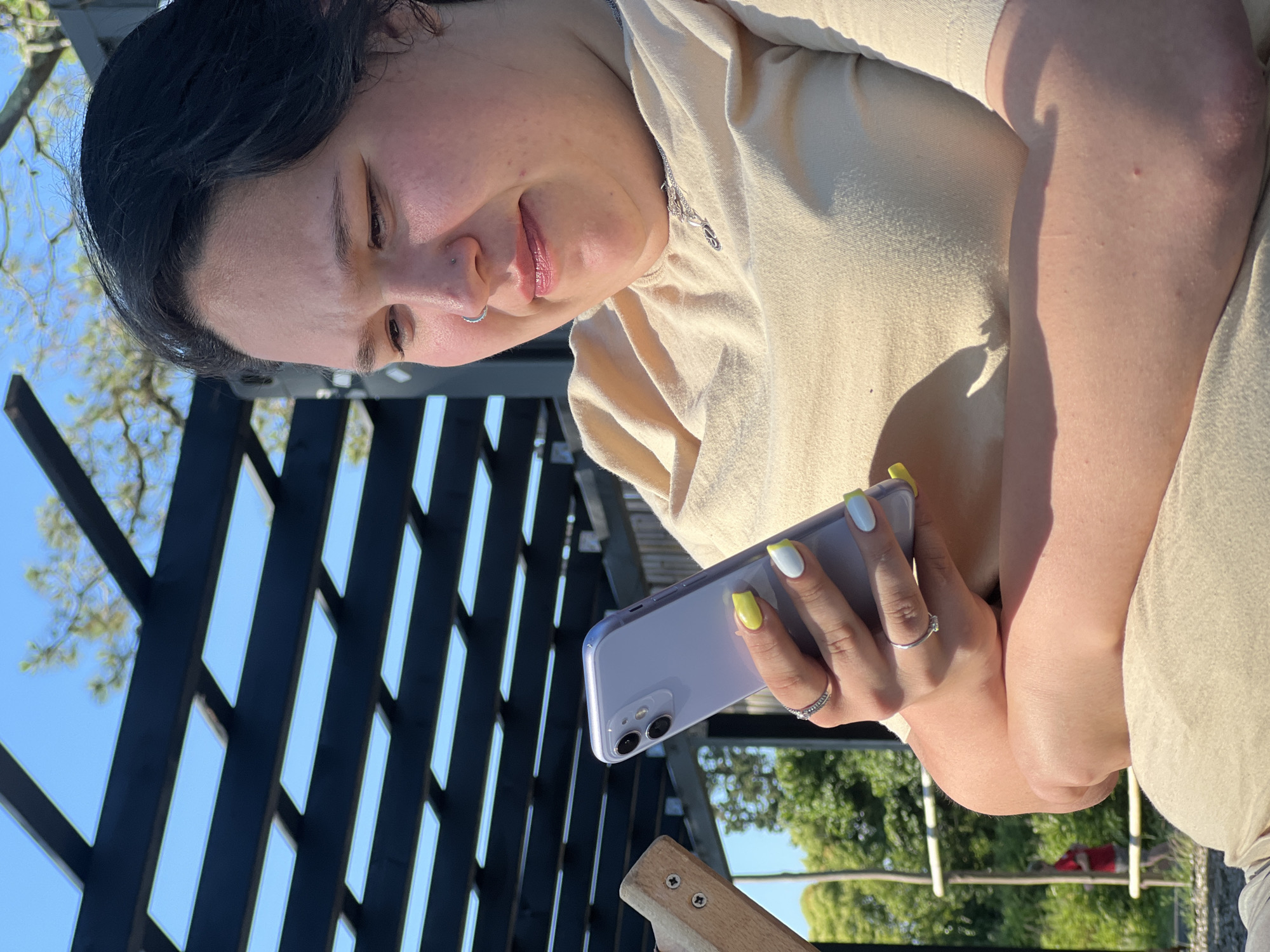 The image shows a young woman outdoors, holding a smartphone. She has dark hair and is wearing a light-colored shirt. She is looking down at her phone with a slight smile. Her nails are painted with a yellow and white design, and she is wearing two rings on her left hand. The background includes a wooden structure, greenery, and a bright blue sky, suggesting it is daytime and the weather is pleasant. The overall mood appears relaxed and casual. The setting is outdoors, possibly a patio or garden area.