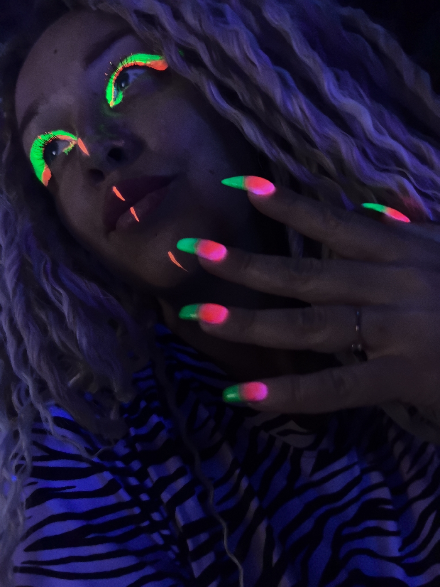 Under blacklight, a person with light-colored locs is facing the camera, partially obscured. The individual's eyes are accentuated with green and orange neon makeup, complemented by corresponding lines on their lips. Their nails are painted with a two-tone design: green tips fading into a pink base. The person's right hand is visible, adorned with a ring on one finger. They wear a zebra-striped shirt that is illuminated in blue by the blacklight. The specific location in the city of Noviy, Russia is not identifiable from the details provided.